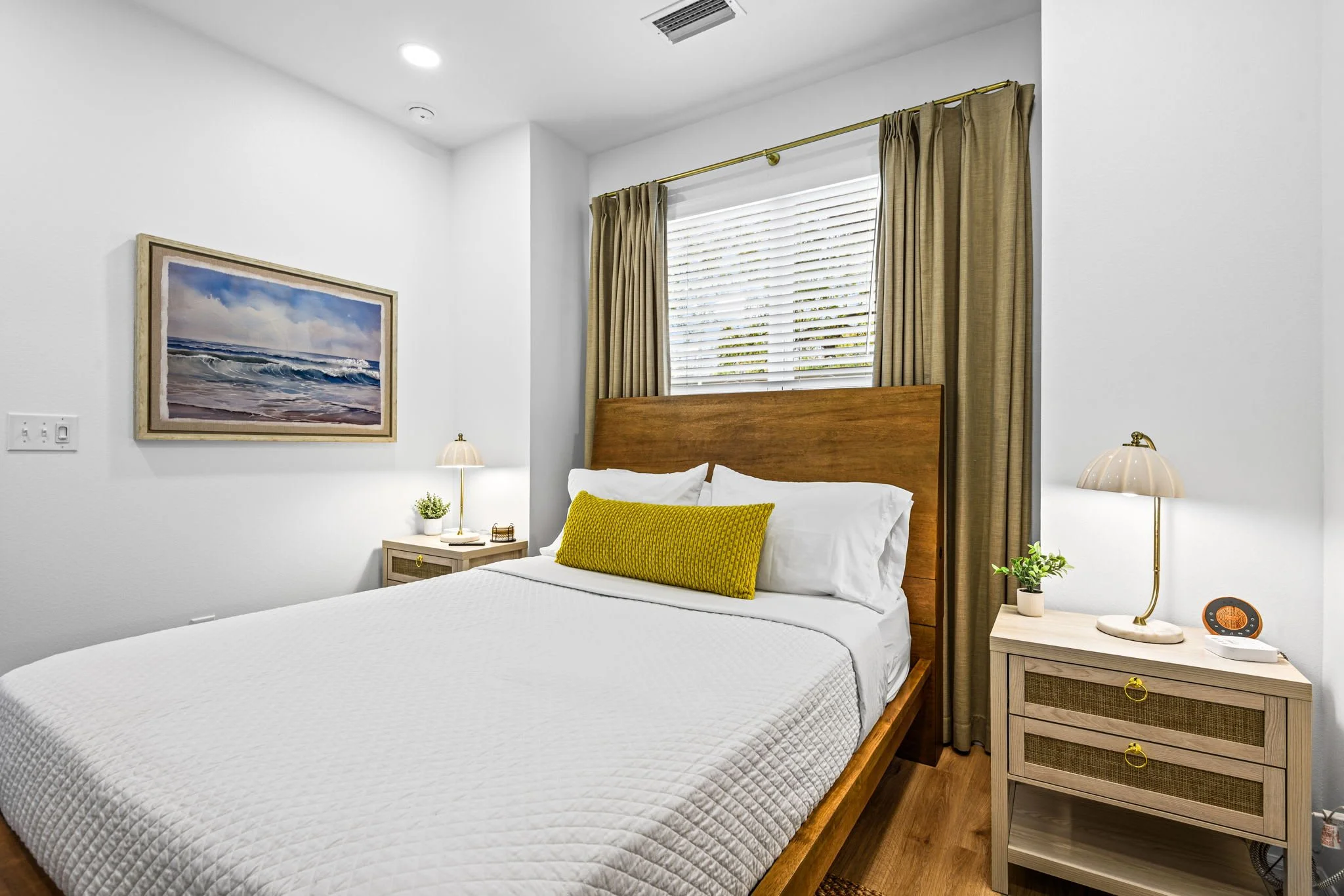 A neatly made bed with white bedding and a long yellow pillow, wooden headboard, situated next to beige nightstands with matching lamps, a painted ocean scene above, and curtains covering a window in a bright, modern bedroom.