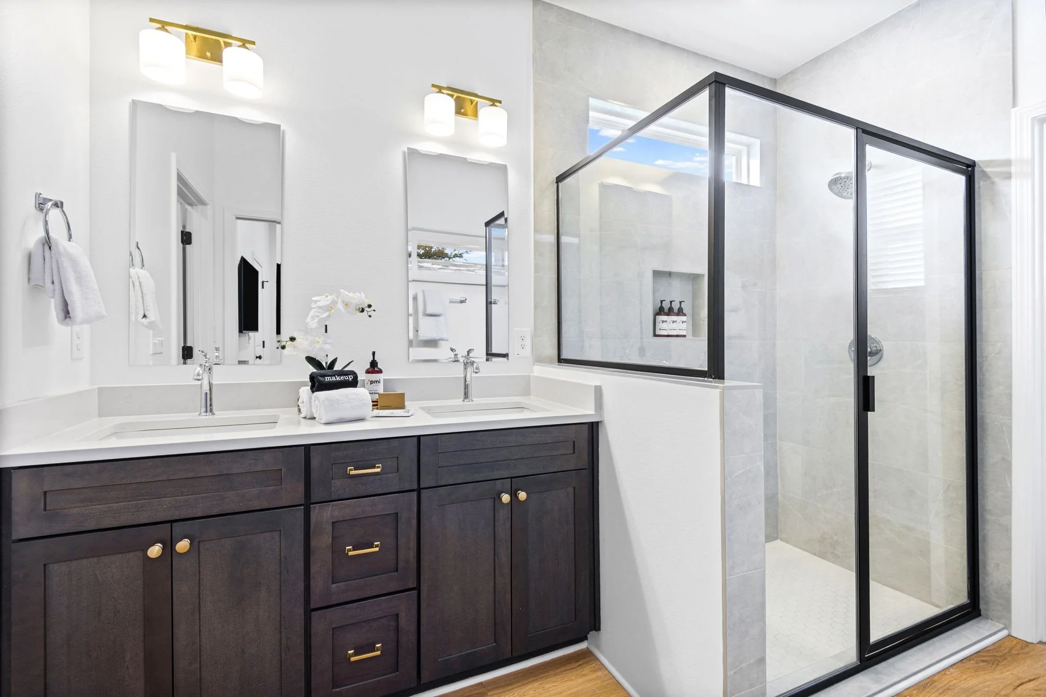 Modern bathroom with dark wood vanity, double sinks, and gold hardware. Large mirror above sinks, wall-mounted lights, white towels, and toiletries on the counter. Glass shower with black frame and tiled interior off to the side.