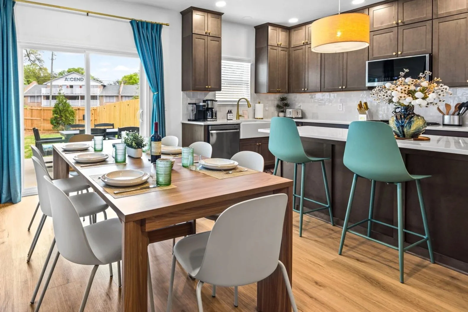 Open concept kitchen and dining area with wooden table set for six, adjacent to a kitchen with dark wooden cabinets, a white counter, bar stools, and a view of a backyard with a wooden fence and outdoor seating.