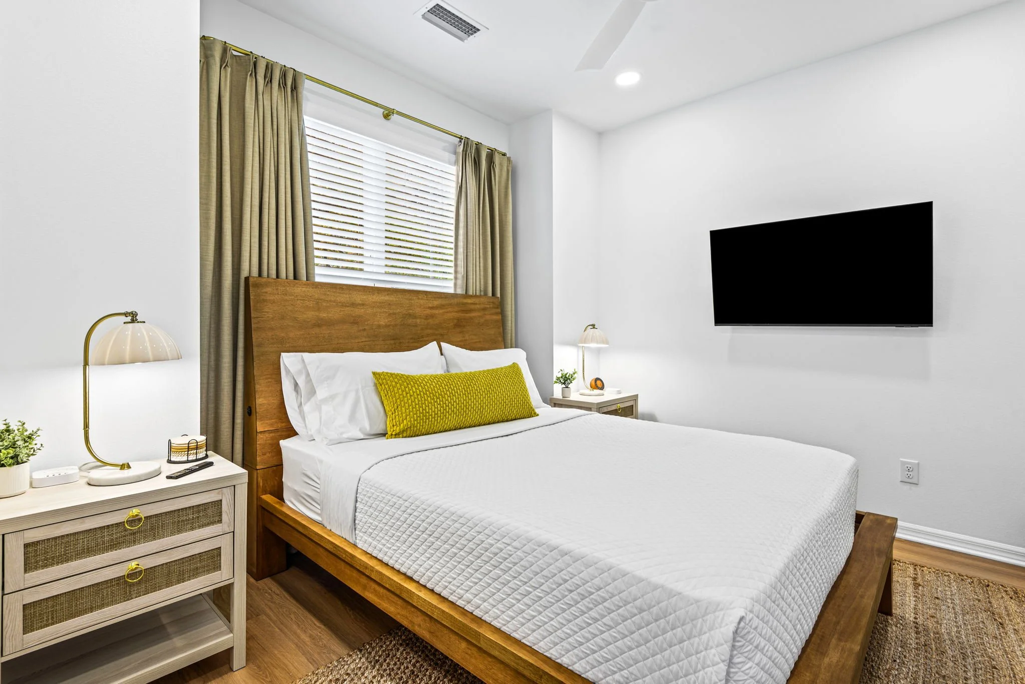 A bedroom with a wooden bed frame, white bedding, and a lime green pillow. There are beige curtains and blinds covering the window, two bedside tables with lamps, a flat-screen TV on the wall, and some small decorative items on the tables.