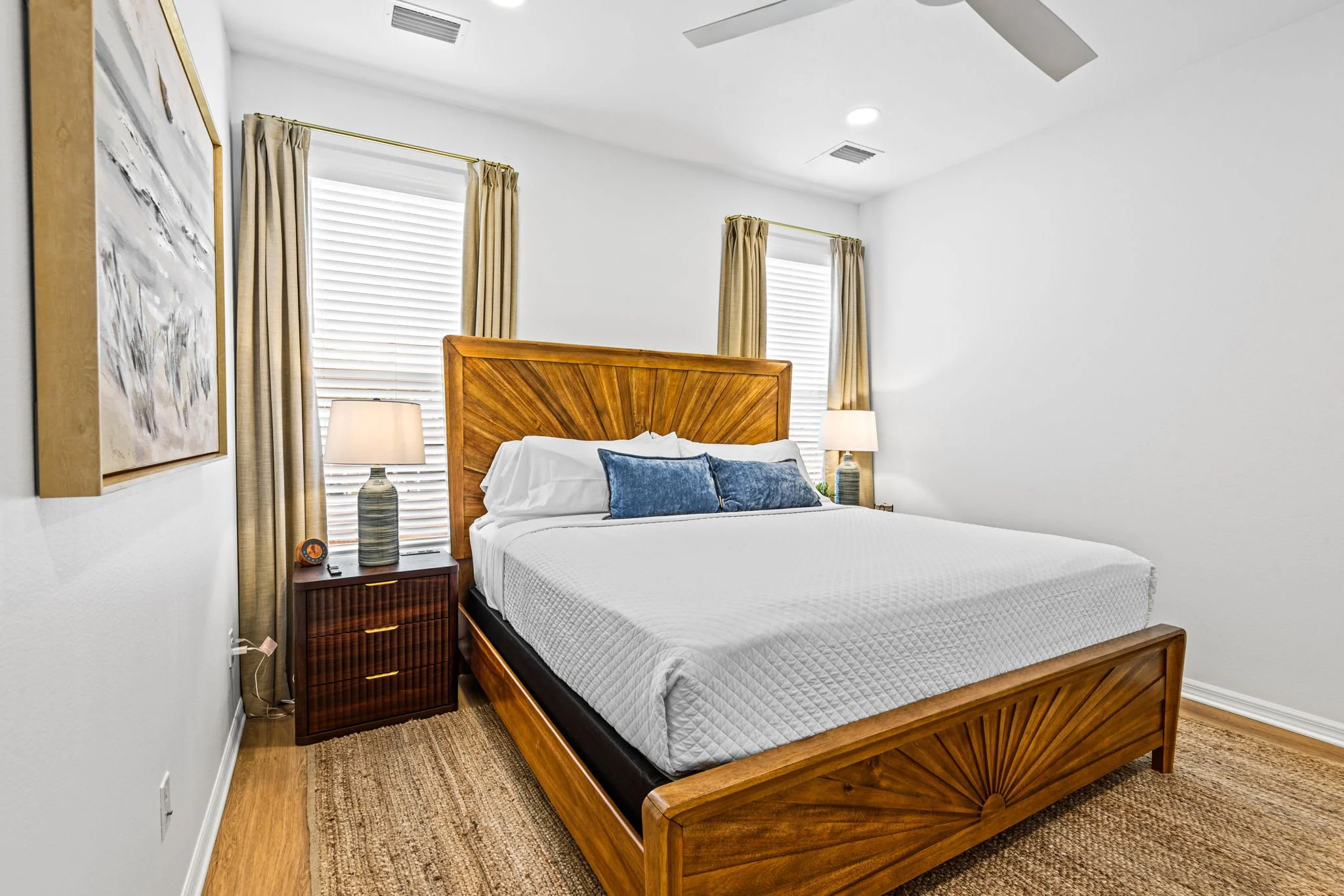 A modern bedroom with a wooden bed frame and white bedding, two blue pillows, two beige curtains, two matching bedside lamps, two windows with blinds, ceiling fan, and wall art.