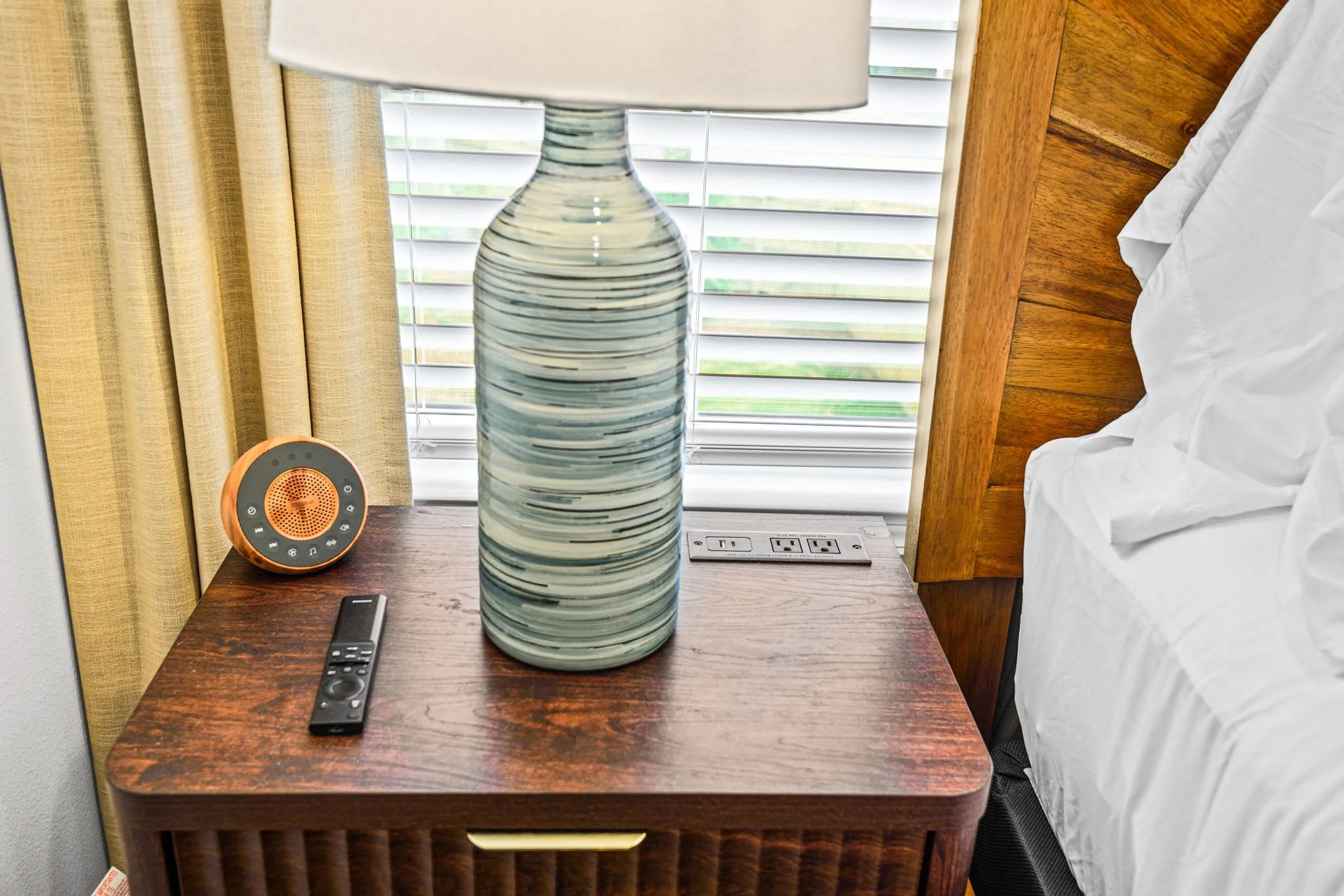 A bedside table with a large striped ceramic lamp, a white noise machine, and extra electrical outlets, with a bed and window with blinds in the background.