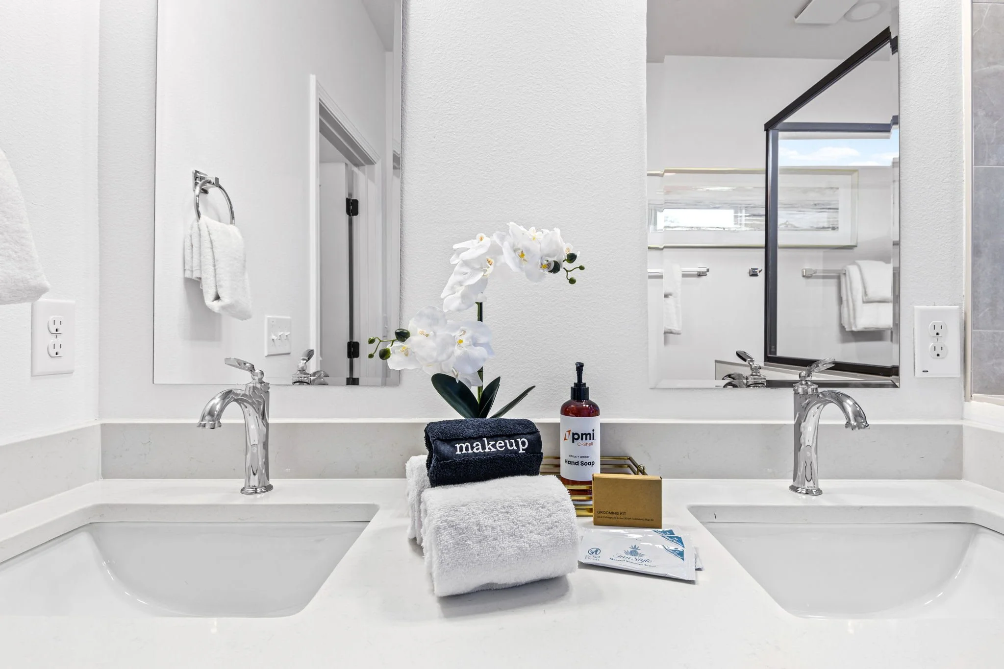 Bathroom vanity with two sinks, topped with toiletries and decorative items, mirror, faucet, orchid flower, hand soap, towels, grooming kit, and brochures, with a window in the background.