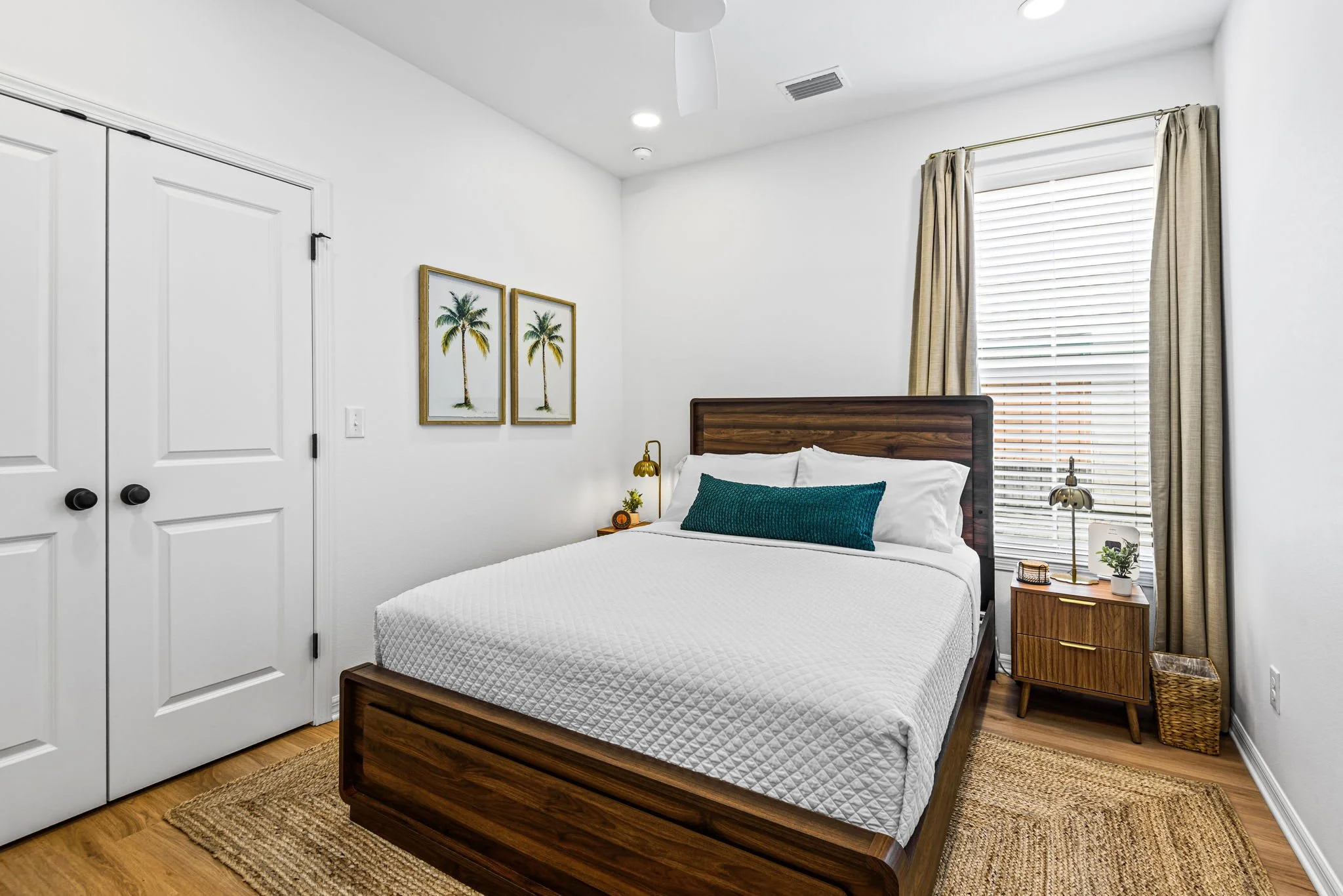 A modern bedroom featuring a wooden bed frame with a white quilted mattress, a green decorative pillow, and a nightstand with a gold lamp, potted plant, and decorative items. There are two framed pictures of palm trees on the white wall, a window with blinds and beige curtains, and a white door with black handles. The floor is wood with a woven rug.