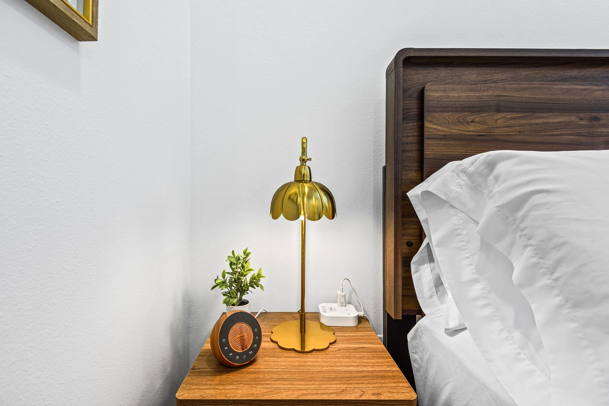 A bedroom nightstand with a gold lampshade, a potted plant, a round digital alarm clock, a white landline phone, and a power outlet with a plugged-in device. Part of a bed with white bedding and a dark wood headboard are visible.