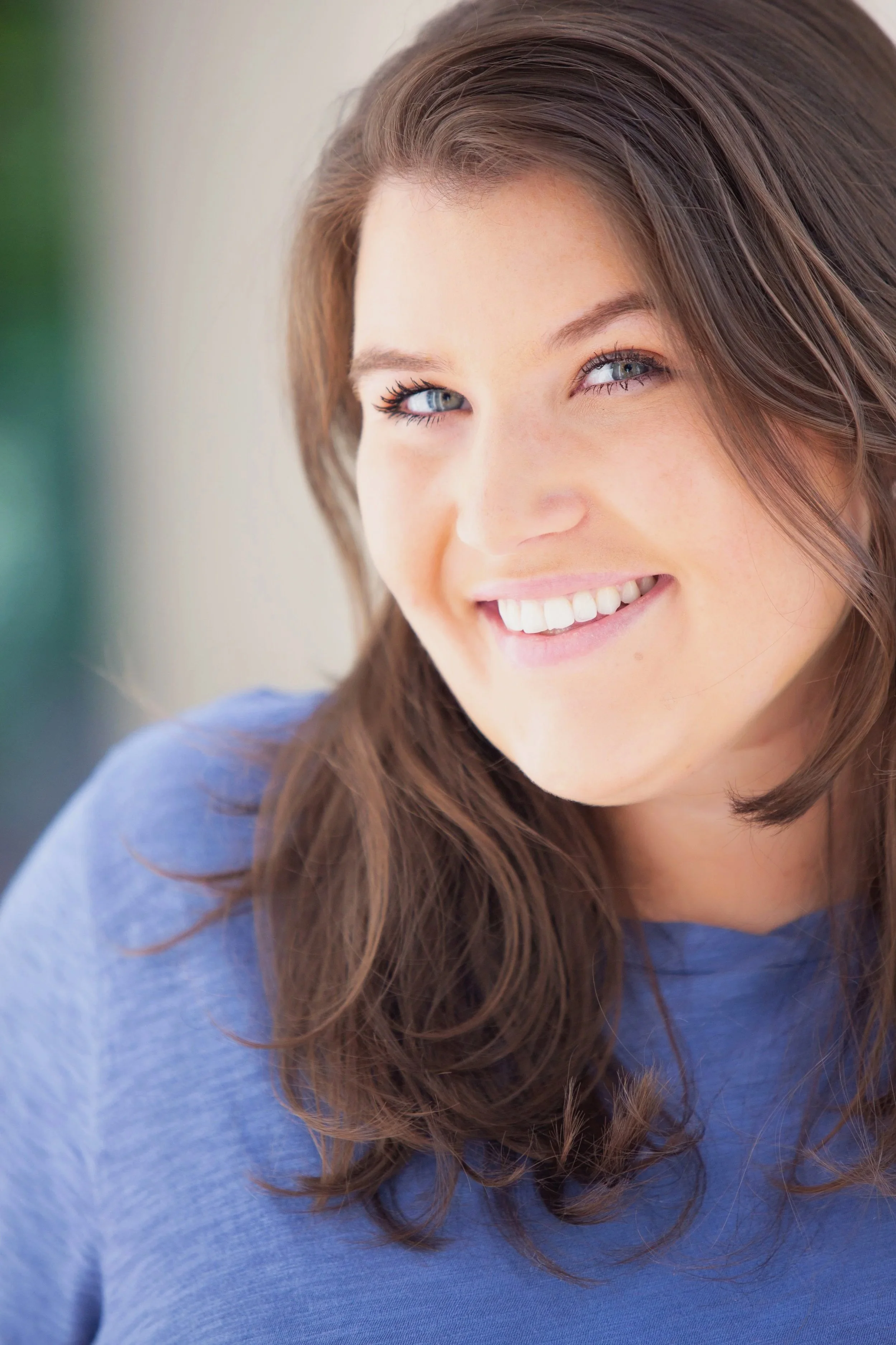 Close-up of a kind, young woman who is a therapist with long brown hair, blue eyes, and a bright smile, wearing a blue top.