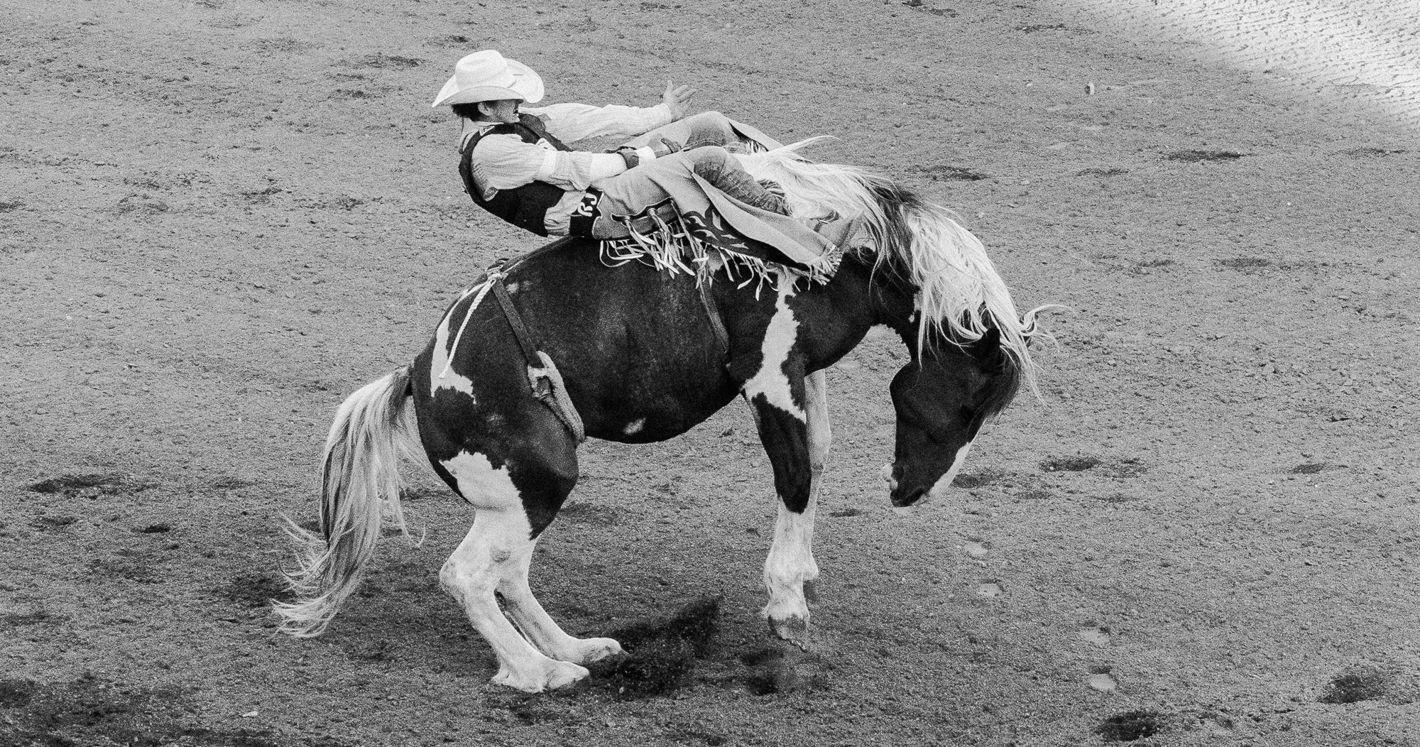 A person riding a horse on a dirt arena, wearing cowboy attire including a cowboy hat, vest, and jeans, with the horse having a black and white coat.