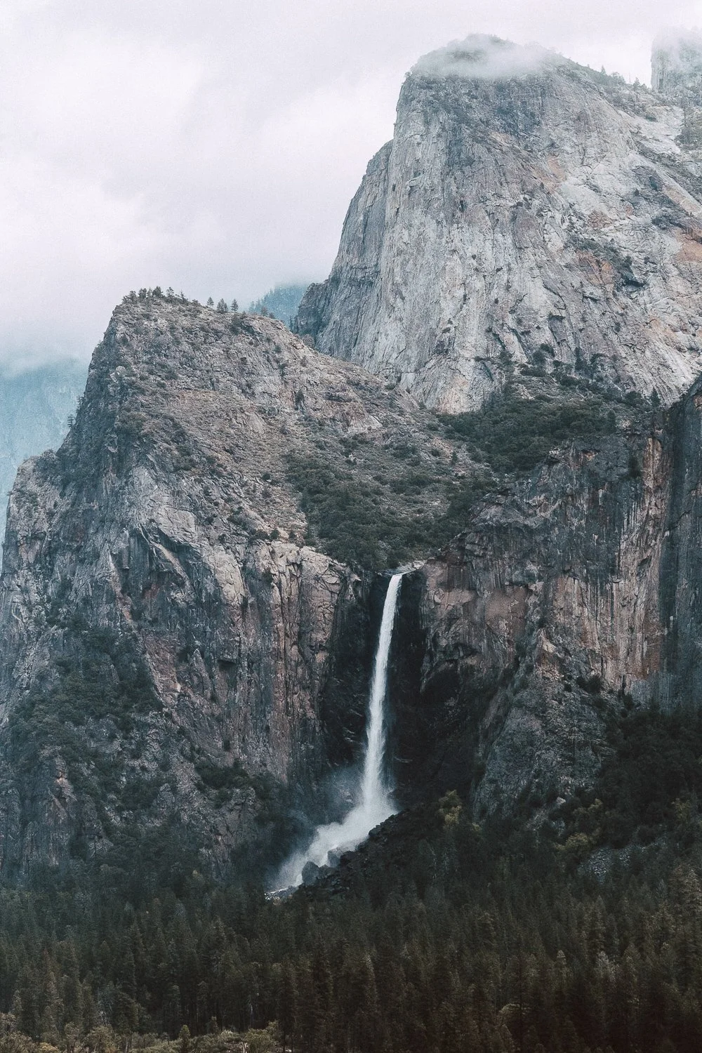 Yosemite National Park, California. A mountain landscape featuring a tall waterfall cascading down a rocky cliff surrounded by trees and mist.