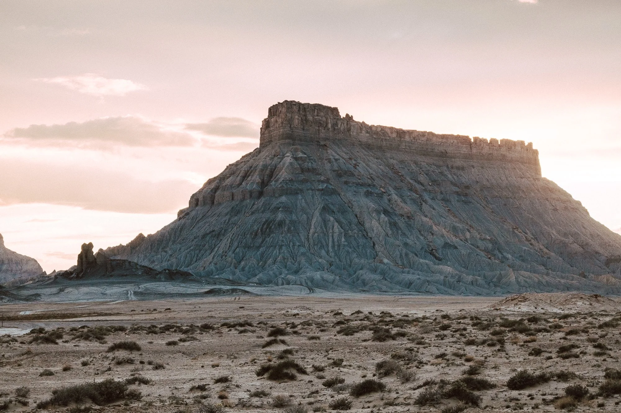 Factory Butte, Hanksville Utah. A large, flat-topped mountain or mesa with sharply sloped sides sits in a barren desert landscape at sunset, with a few scattered bushes in the foreground.