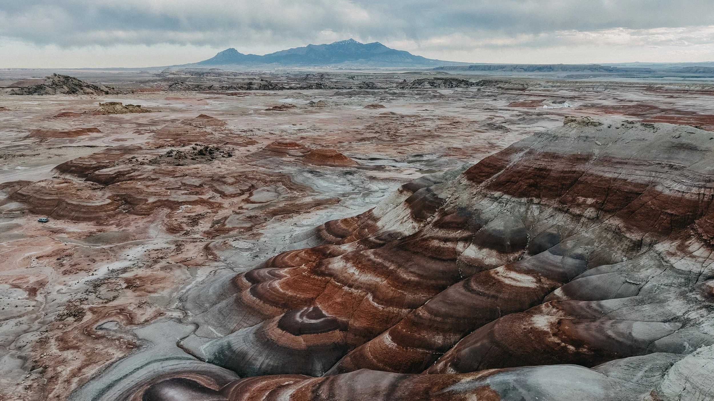 A vast desert landscape with colorful, layered rock formations and distant mountains under a cloudy sky.