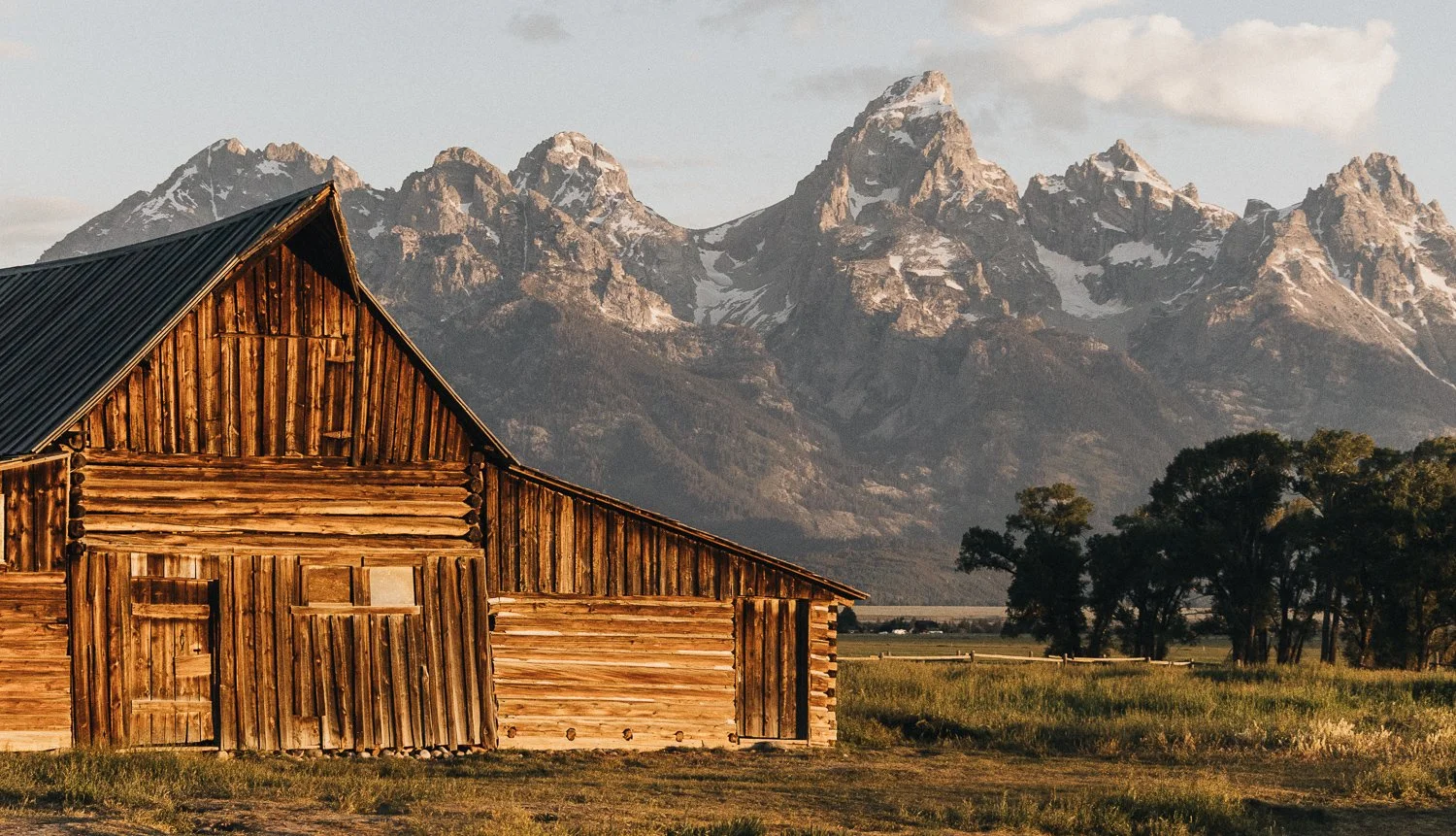 Grand Teton National Park the Moulton Barn. A rustic wooden barn with a metal roof, set against a backdrop of snow-capped mountains and green trees in a rural landscape.