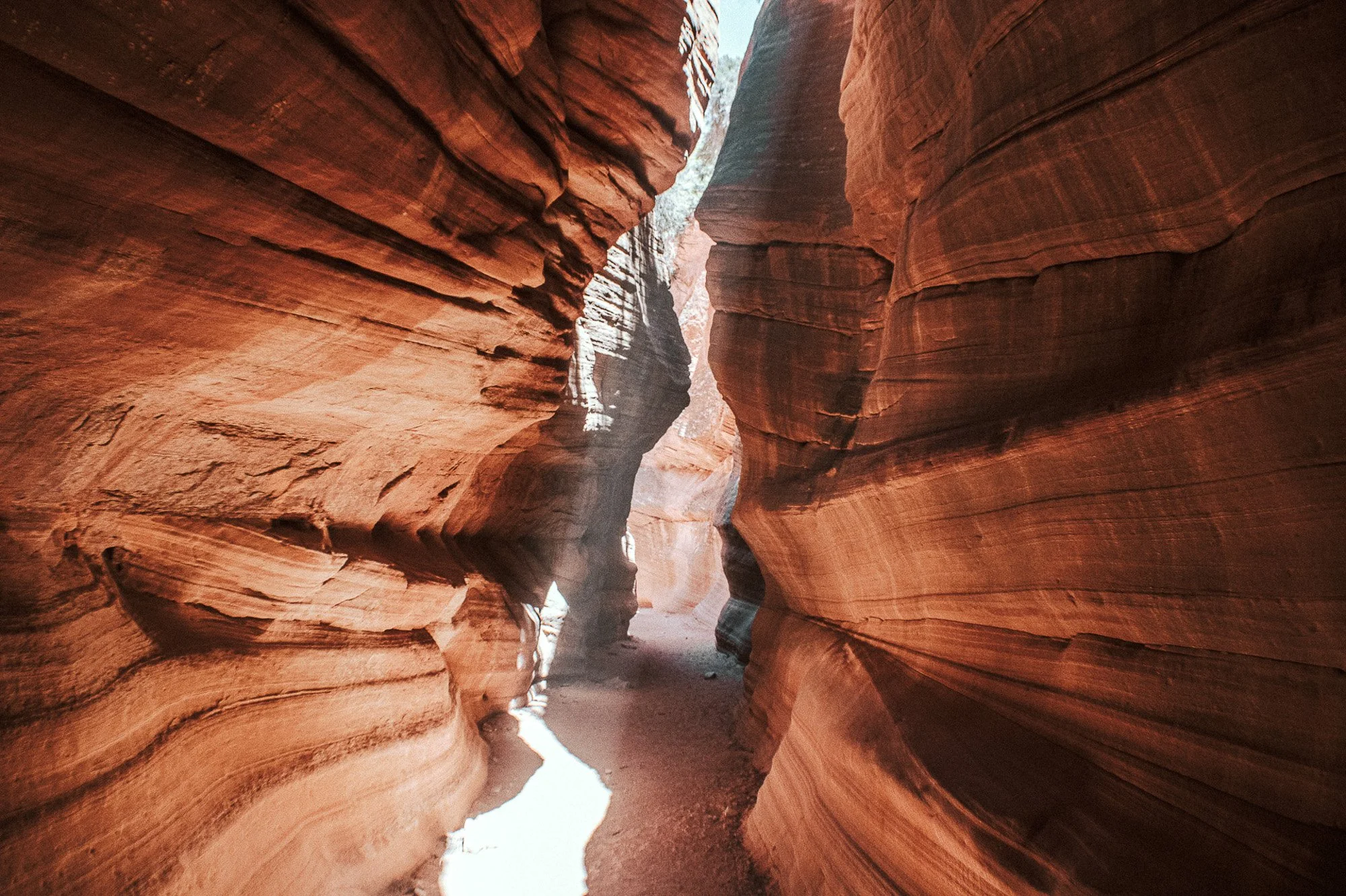A narrow slot canyon with smooth, layered red and brown rock walls, with sunlight illuminating parts of the crevices.