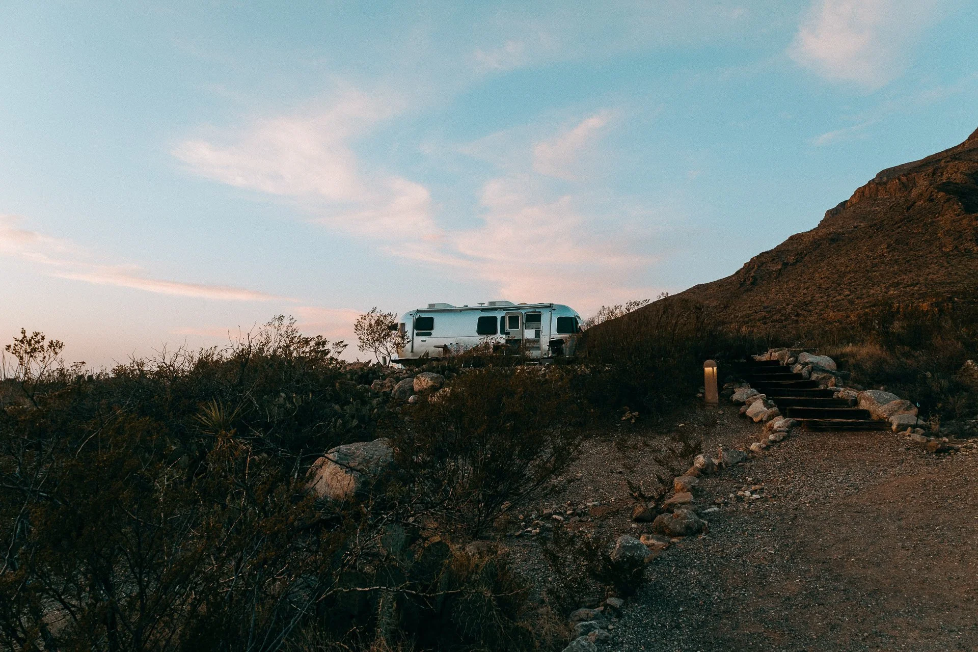 An Airstream parked on a rocky hillside with steps leading up to it, desert shrubs in the foreground, a mountain on the right, and a clear sky with scattered clouds at dusk in the background.