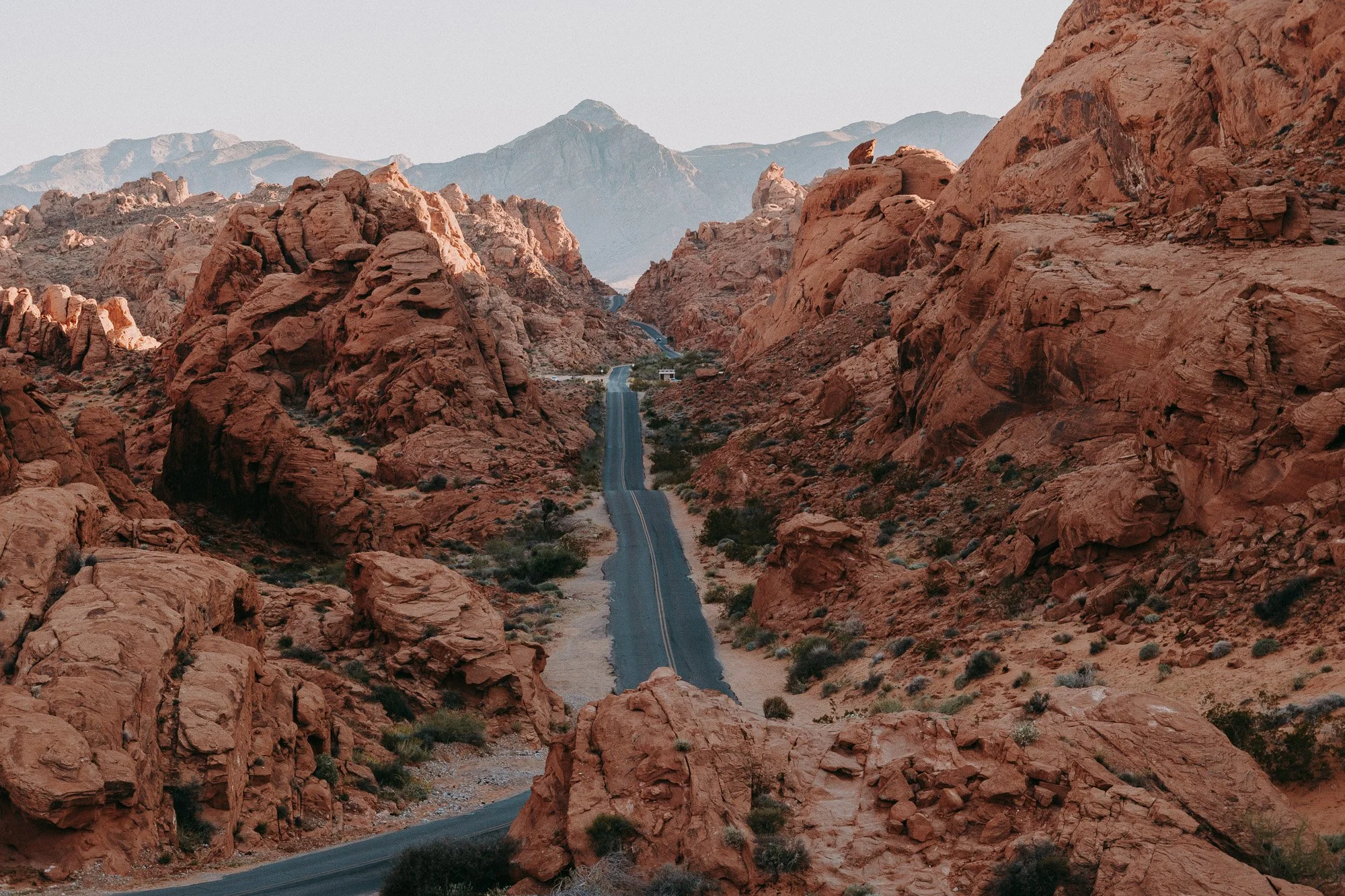 Valley of Fire National Park, Mouse's Tank road. A winding two-lane road through a rocky desert landscape with red sandstone formations and mountains in the background.