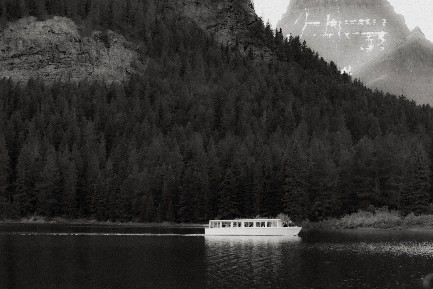 A white passenger boat in Glacier National Park, sailing on a calm lake with a dense forest and mountain cliffs in the background.