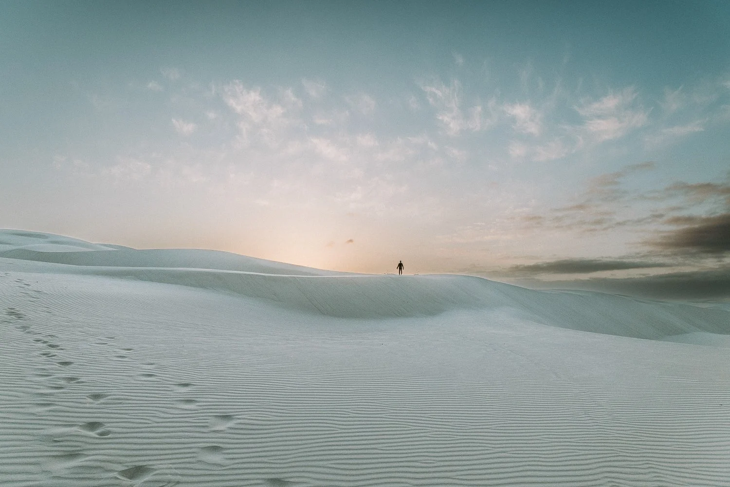 A person standing alone on a vast, white California sand dune under a cloudy sky with the sun near the horizon.