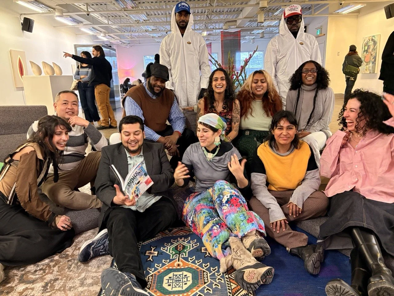 Group of diverse people laughing and smiling in an indoor art gallery, some seated on the floor and others standing behind, with artwork and artwork displays on the walls.