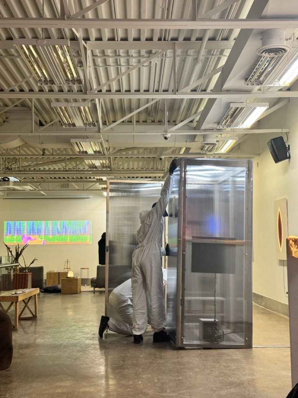Two people in white coveralls working inside a small, transparent enclosure in a modern indoor space, possibly a gallery or event area, with furniture and a colorful digital display in the background.