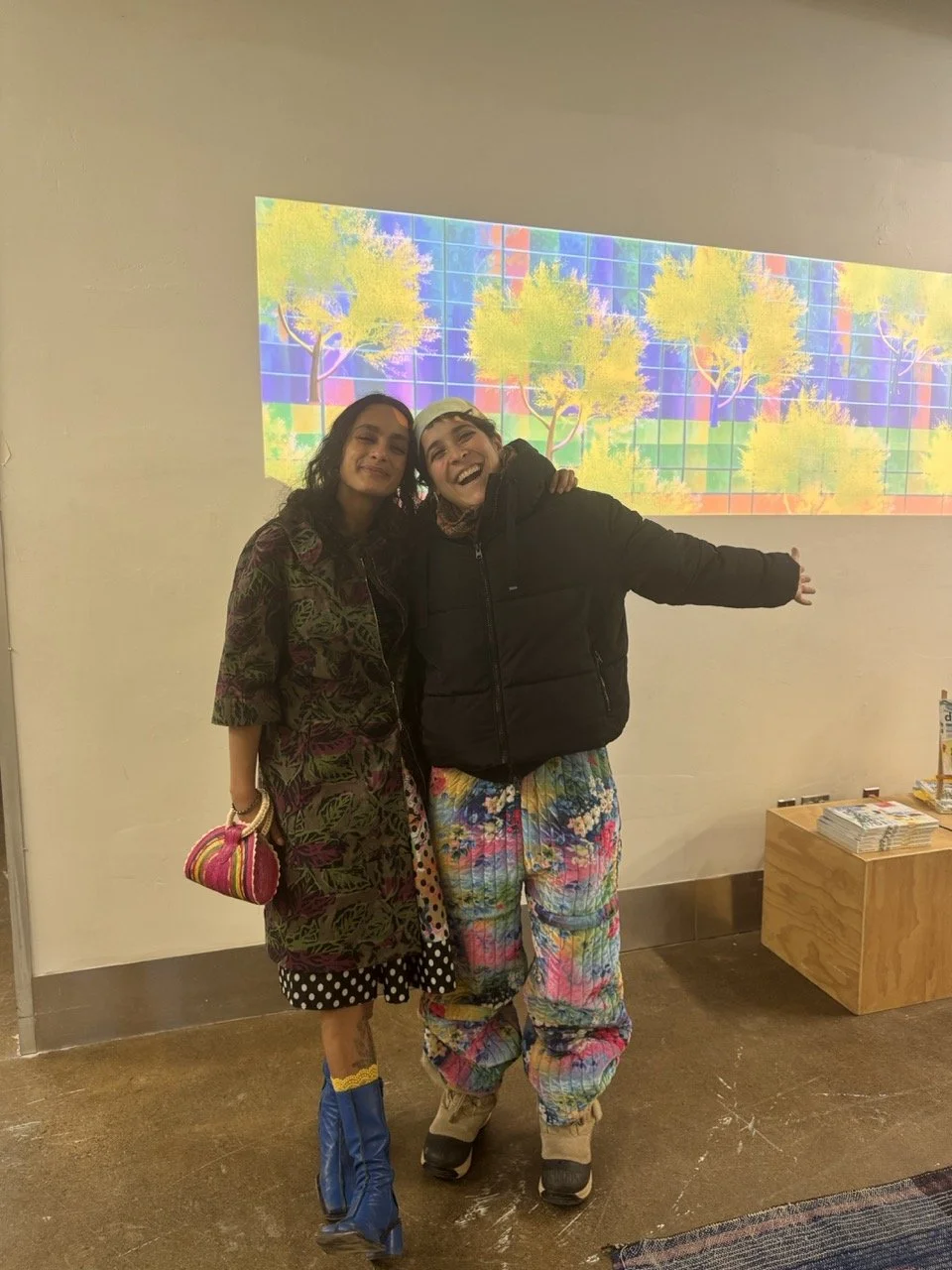 Two women standing together indoors, smiling, with an abstract colorful digital art projection of trees and squares on the wall behind them.