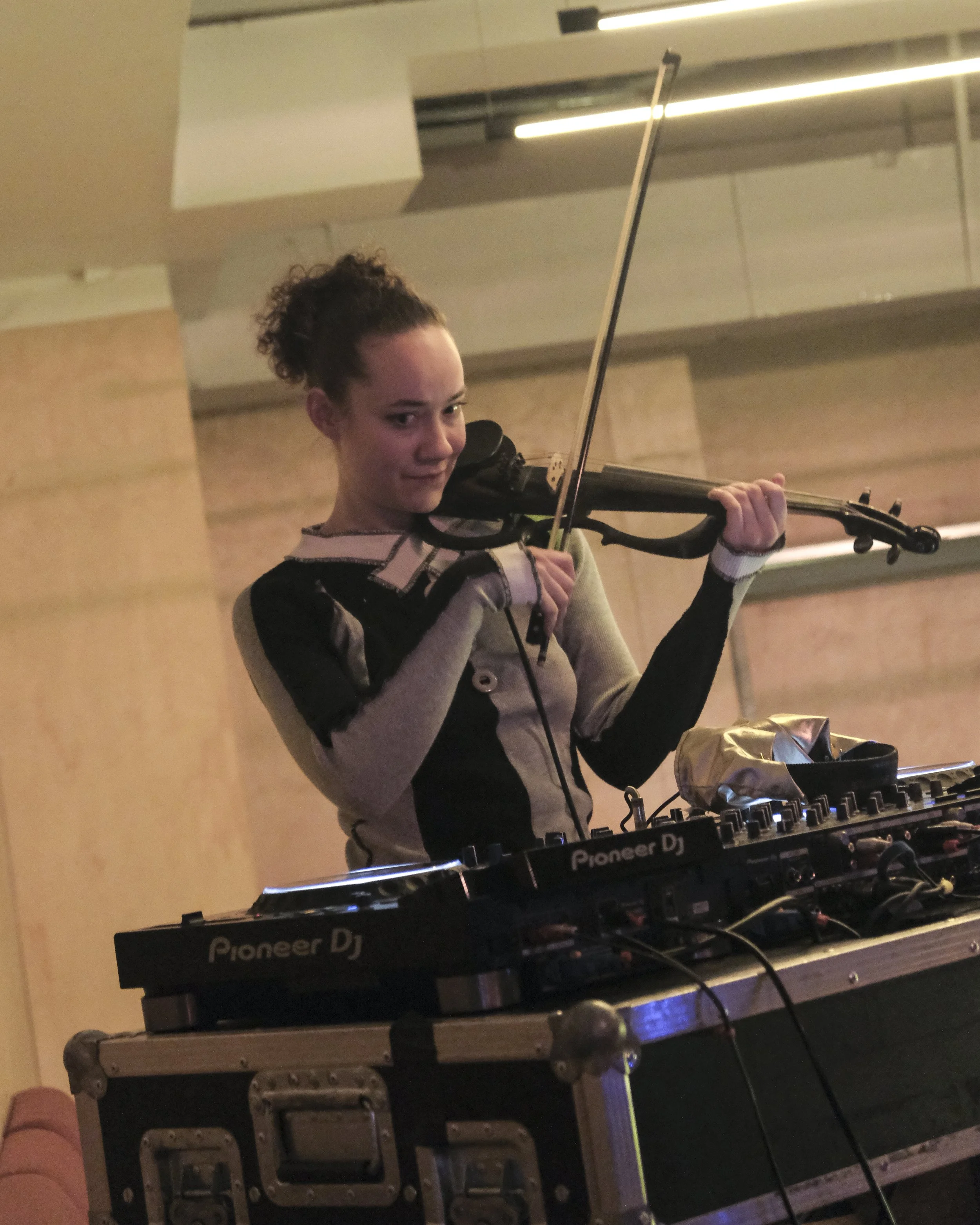 A woman playing a violin in a room with wooden walls and a ceiling-mounted light fixture, positioned behind a DJ setup with Pioneer DJ equipment on a wooden table.
