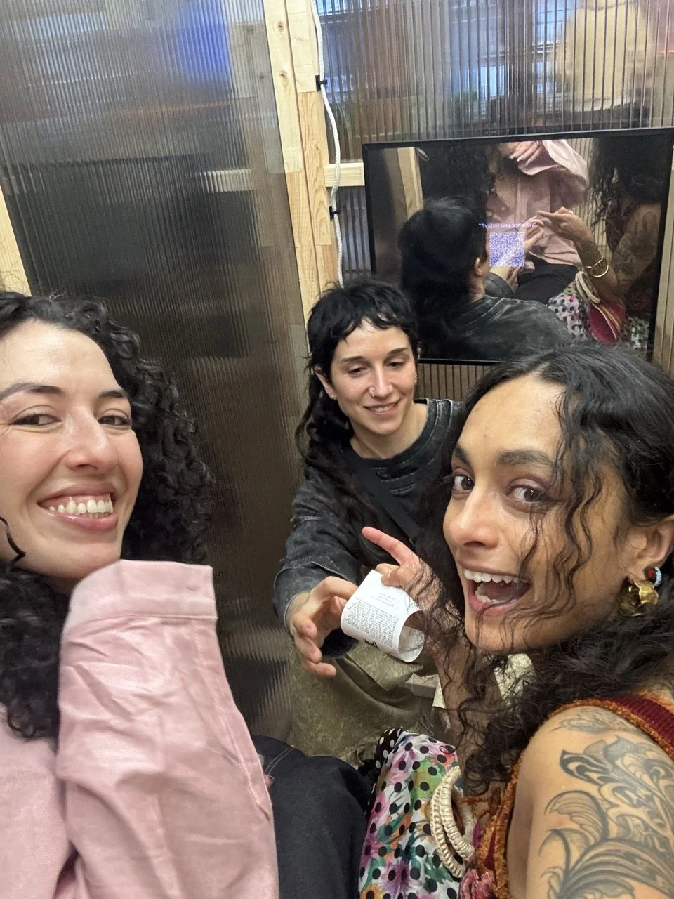 Three women with curly hair smiling and posing for a selfie indoors, with a woman on the right showing a tattoo on her arm and wearing colorful earrings.