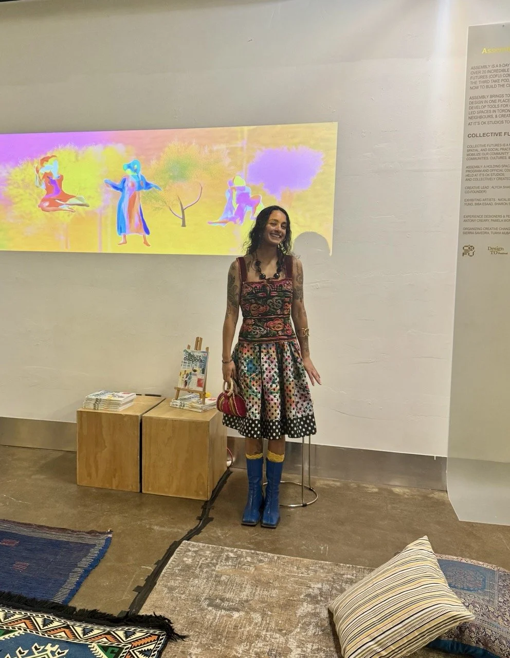 A woman with long dark hair, wearing a colorful embroidered dress and blue knee-high boots, stands smiling in front of a light-colored wall with a digital artwork projected behind her. There are two small wooden tables beside her with magazines or bo