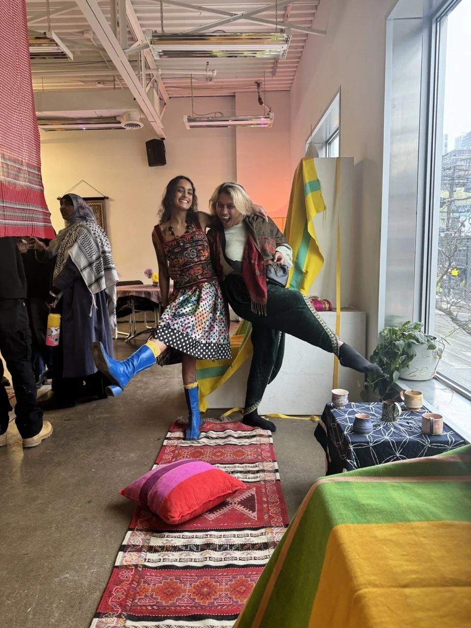 Two women posing playfully with their legs raised, standing indoors near a window and colorful decorations, with a table displaying pottery and a small table with a green and yellow tablecloth in the foreground.