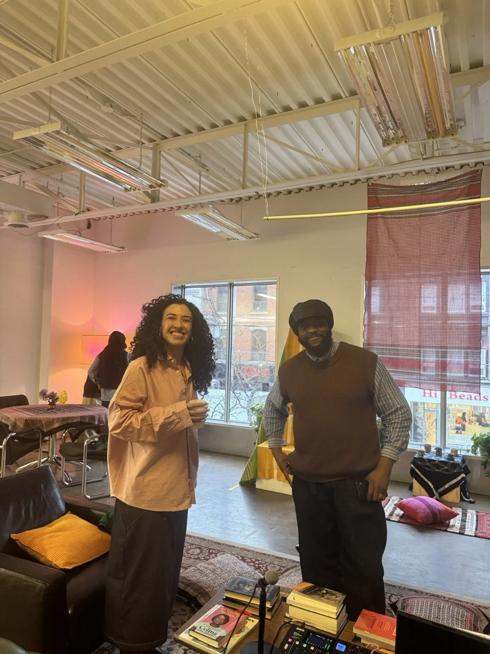 A woman with curly hair and a man wearing a hat and sweater standing inside a cozy, well-lit room with books and a microphone on a table, smiling at the camera.