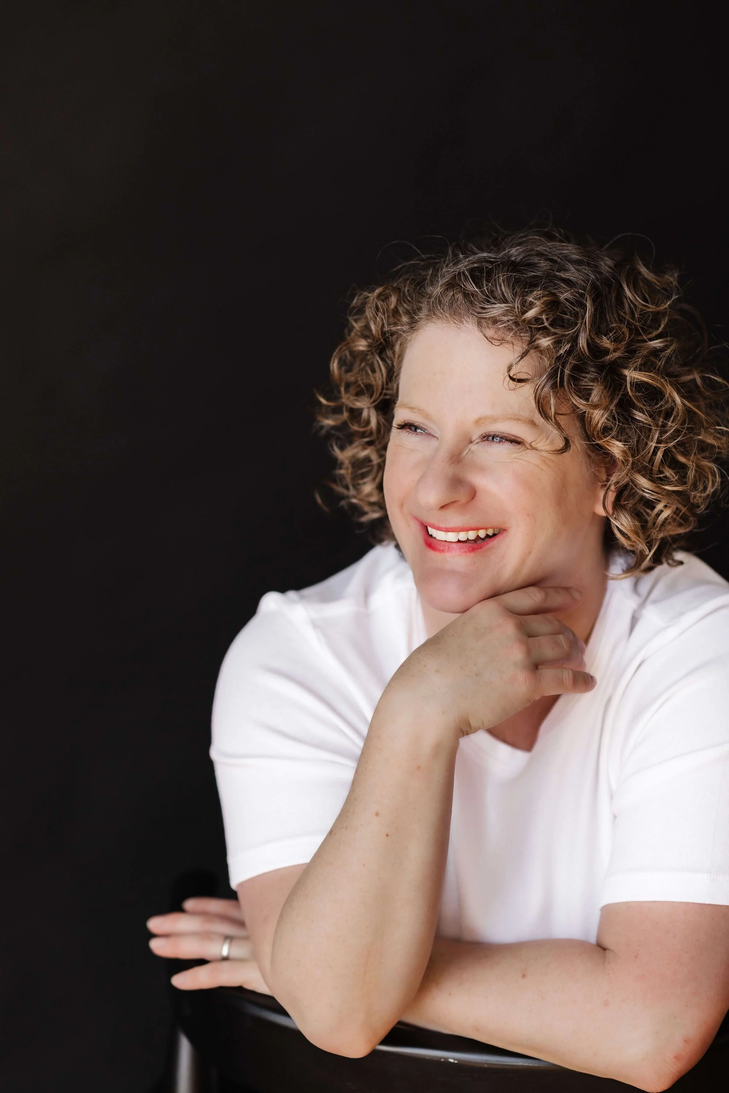 A woman with curly hair smiling, wearing a white shirt, resting her chin on her hand, against a black background.