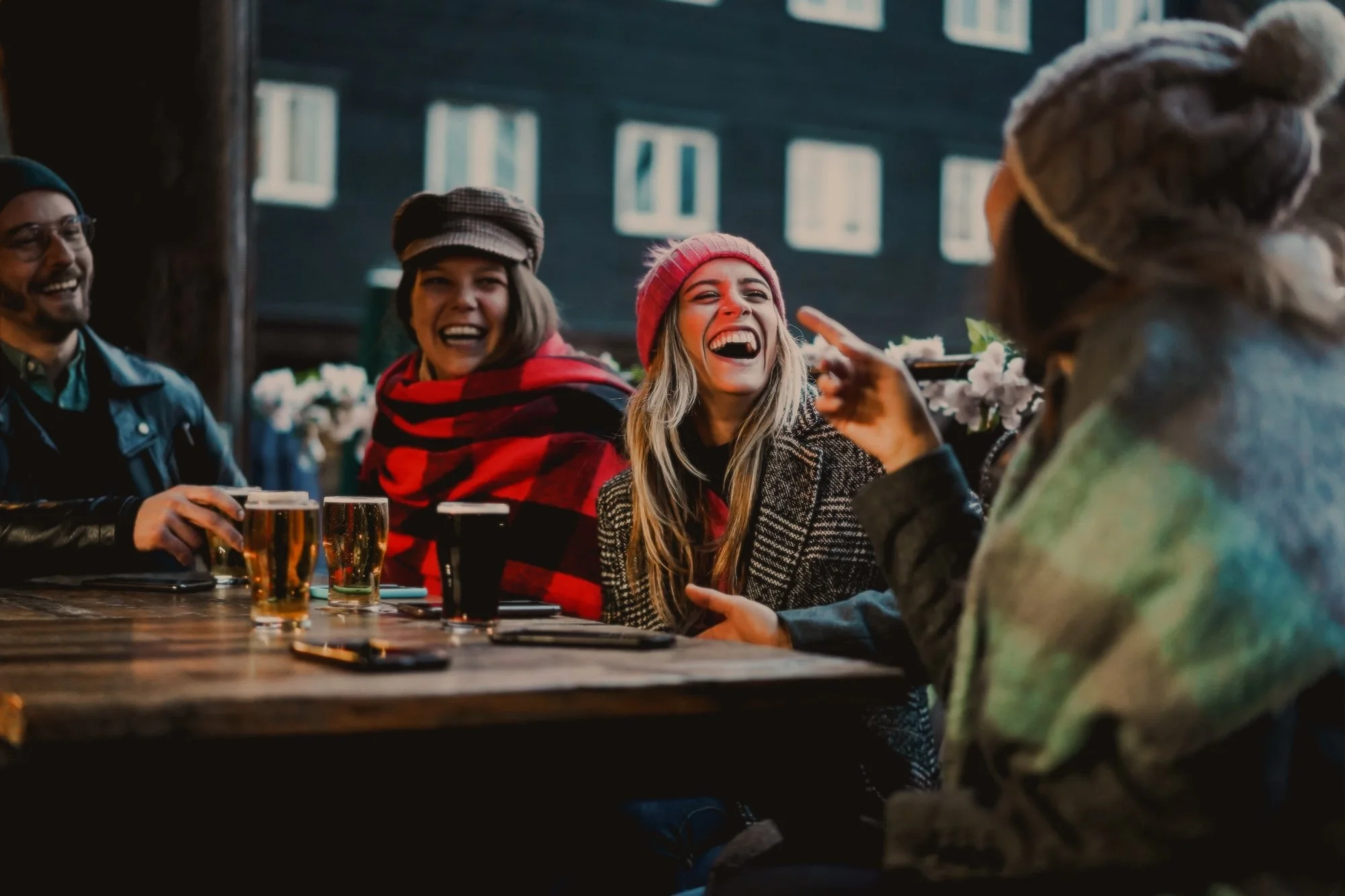 A group of five friends sitting at a wooden table outdoors, laughing and talking. They have drinks in front of them and are wearing winter clothing such as hats and jackets. The scene appears to be set in a cozy, social environment.