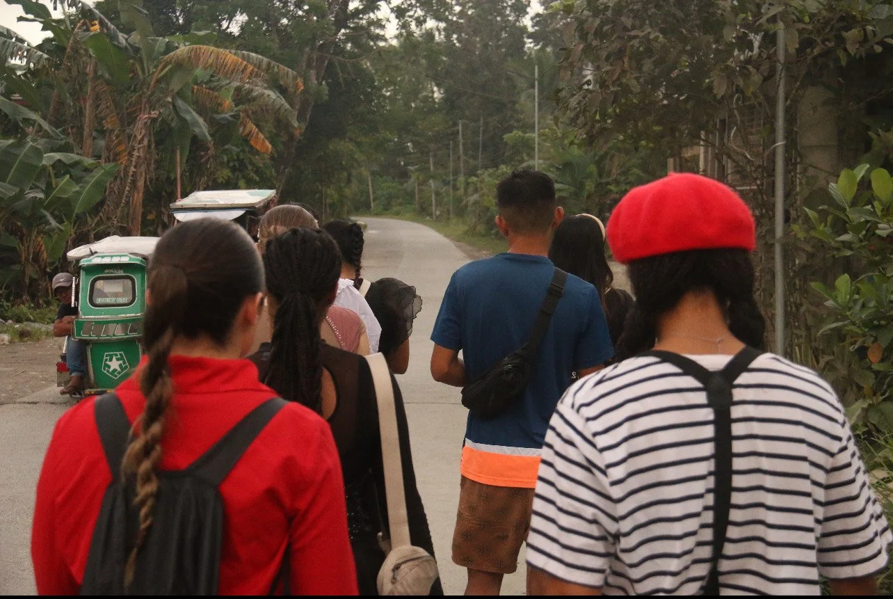 A group of people standing on a rural street with banana trees and greenery on both sides, some with backpacks and hats, waiting or walking together.