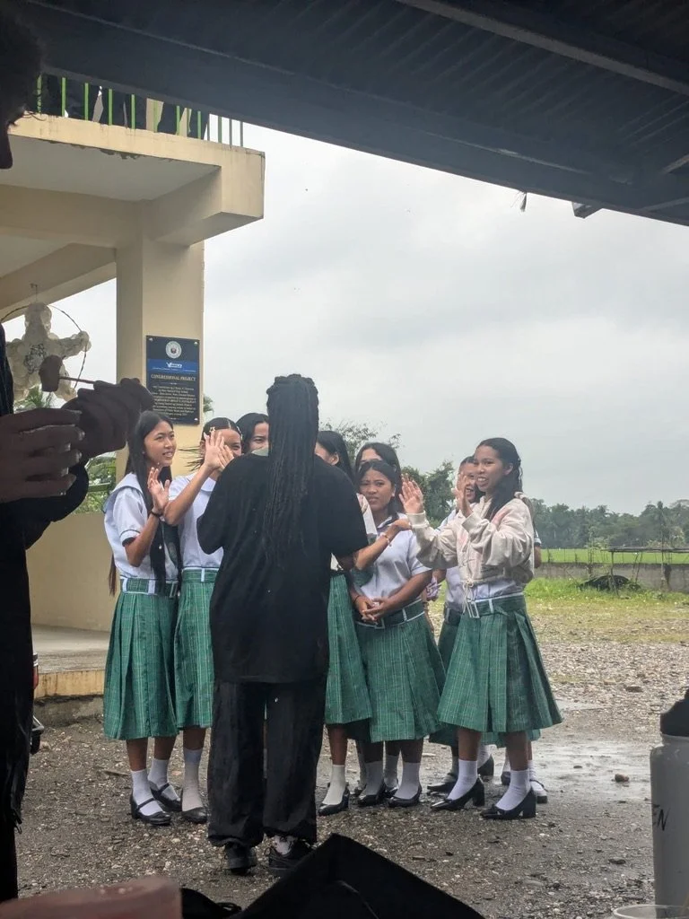 Group of young women in green plaid skirts, white blouses, and socks, standing outdoors under a roof, greeting each other with high fives and handshakes.