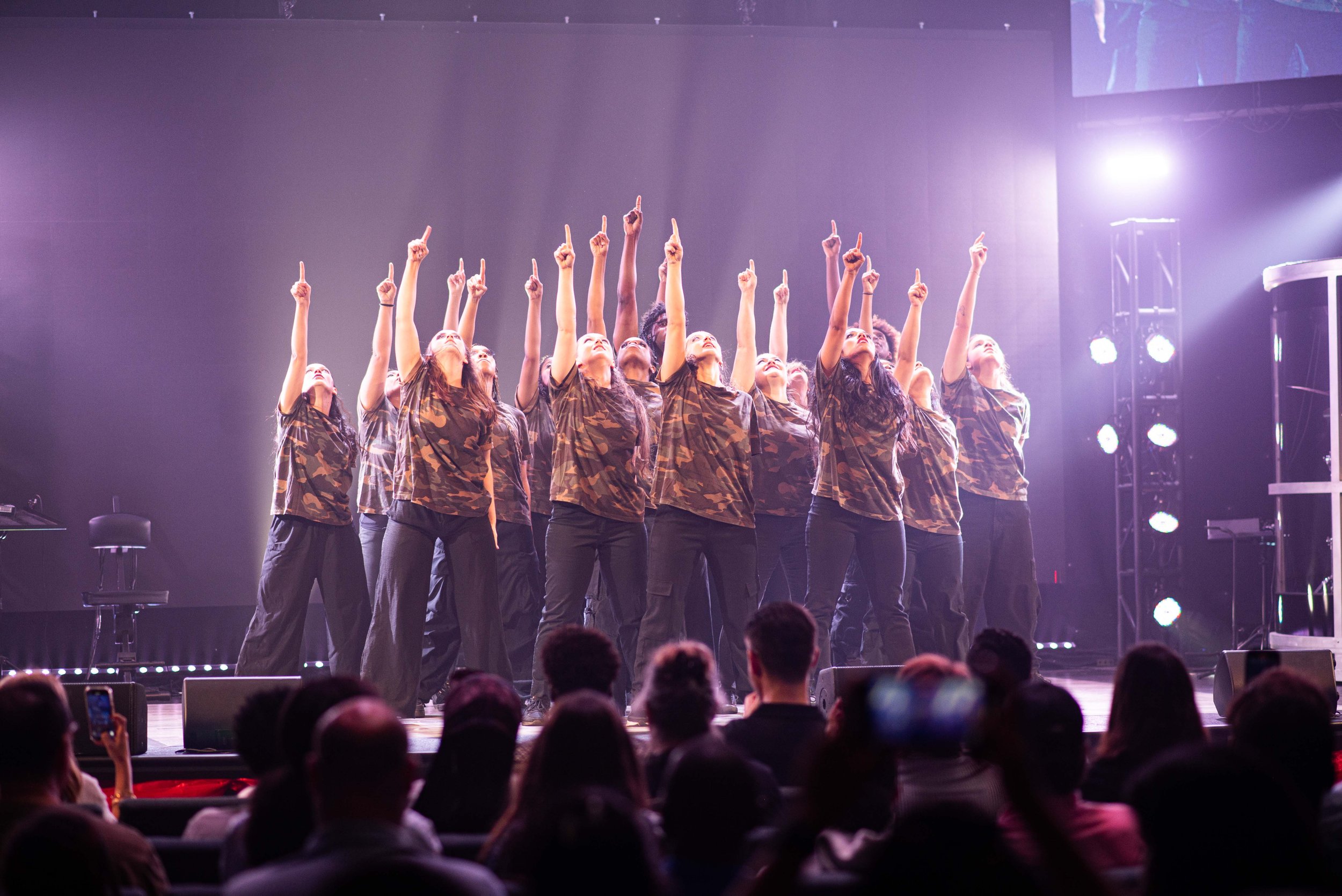 Group of dancers on stage raising one finger each, wearing camouflage t-shirts and black pants, in front of a dark backdrop with stage lights.