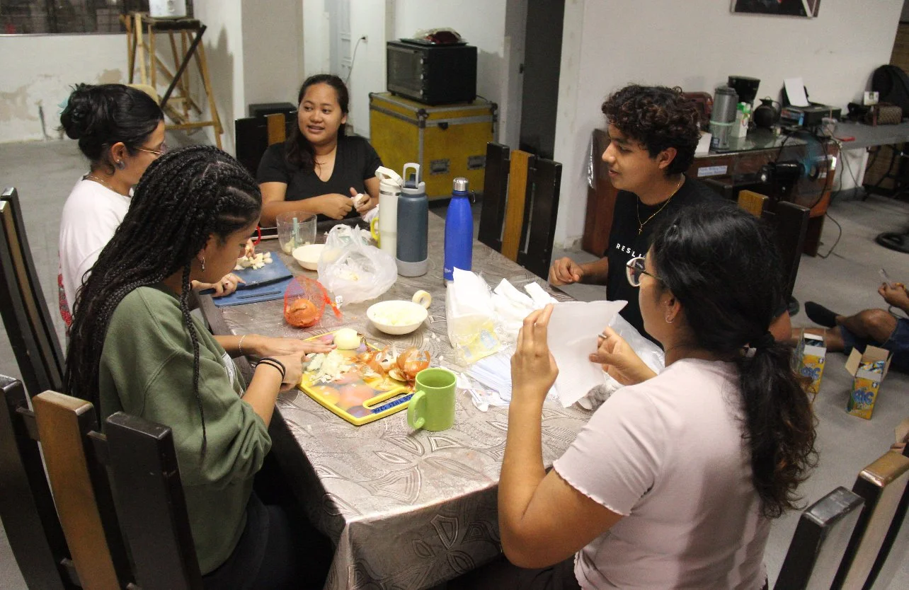 Five women are gathered around a dining table making food, with various kitchen items and ingredients on the table.
