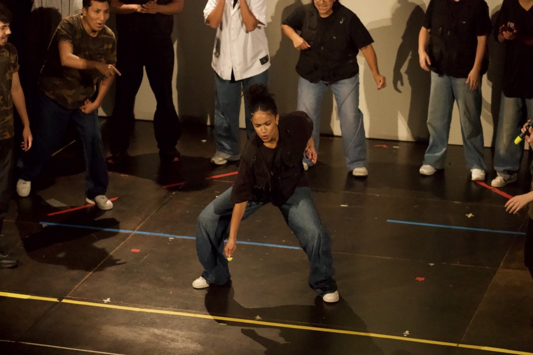 A woman in black shirt and jeans performing a dance move on a black stage, surrounded by a group of people watching.