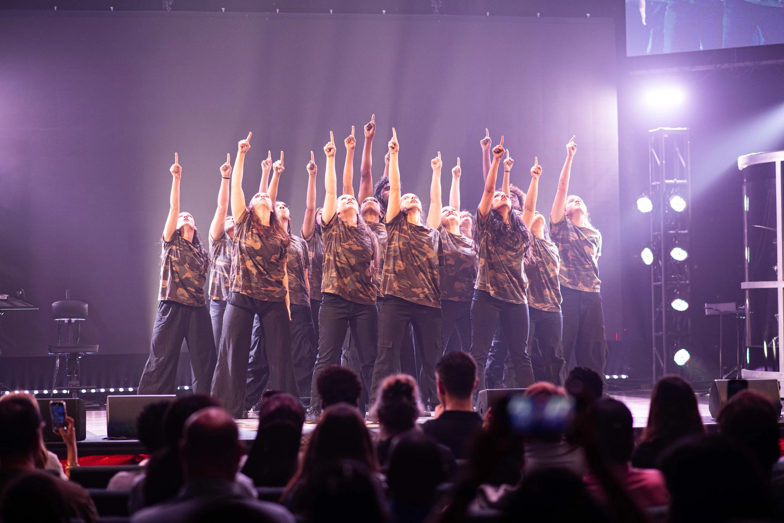 A group of dancers on stage wearing camouflage t-shirts and black pants, pointing upward with one finger, illuminated by stage lights, with an audience watching.