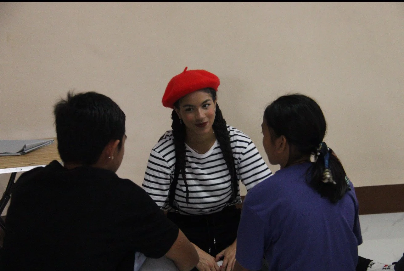 Three young people sitting at a table having a conversation, with a woman in a red beret and striped shirt in focus.