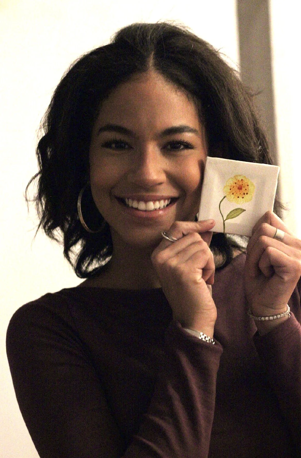 A woman with dark, curly hair smiling and holding a small, white cloth with a yellow flower illustration up to her face.
