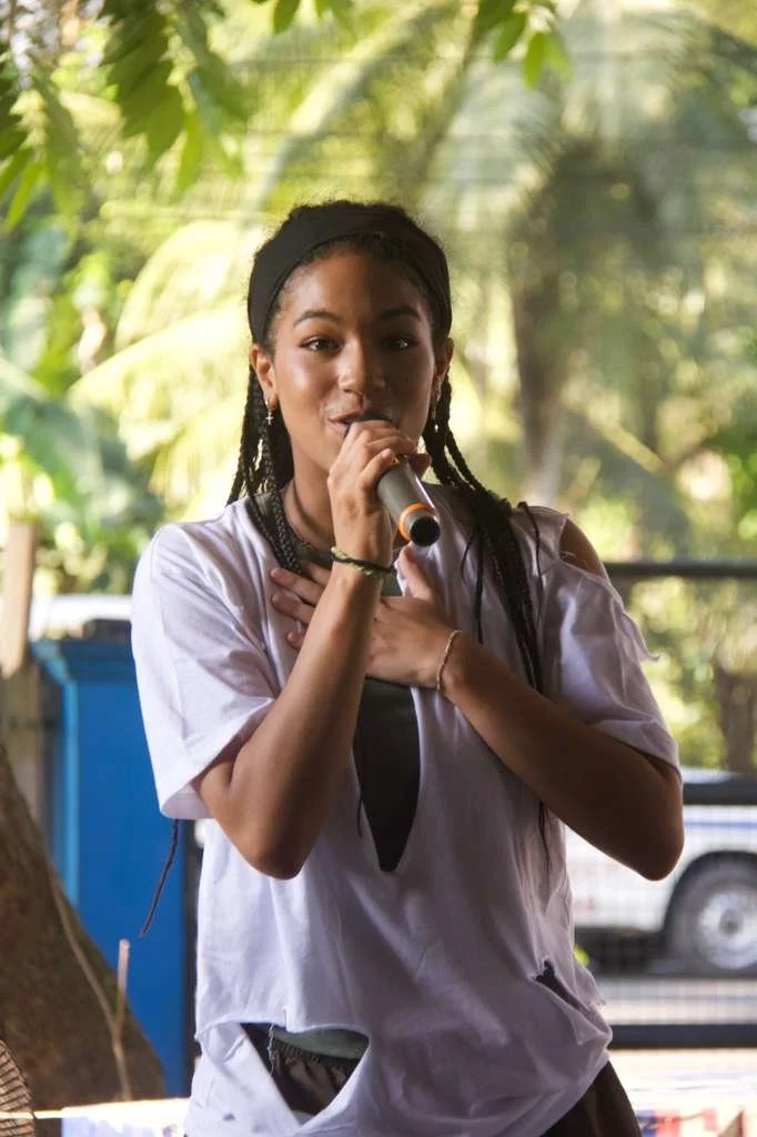 A young woman with braids and a black headband singing into a microphone outdoors with green trees in the background.