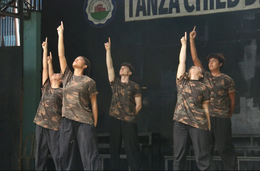 Five young performers dressed in camouflage t-shirts and black pants on stage, raising one arm and gazing upwards, with a banner and logo in the background.