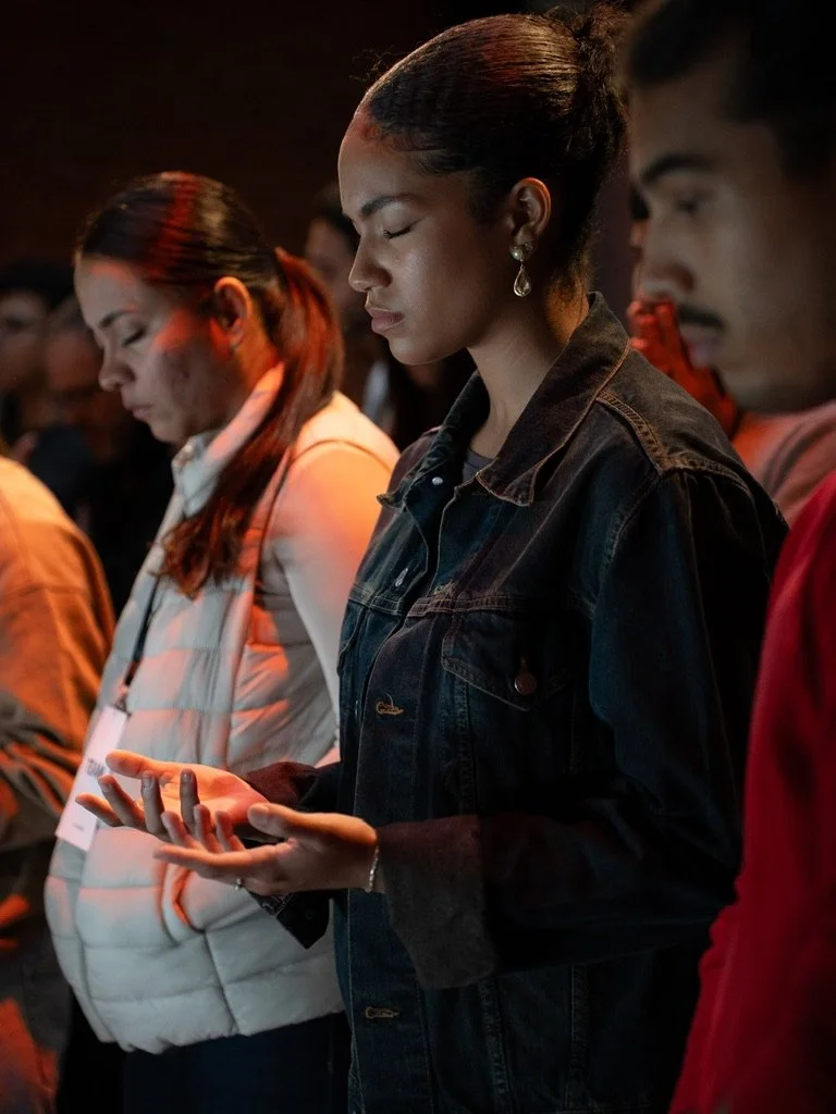 A group of people standing with eyes closed, some holding their hands in a prayer-like position, participating in a moment of contemplation or prayer.