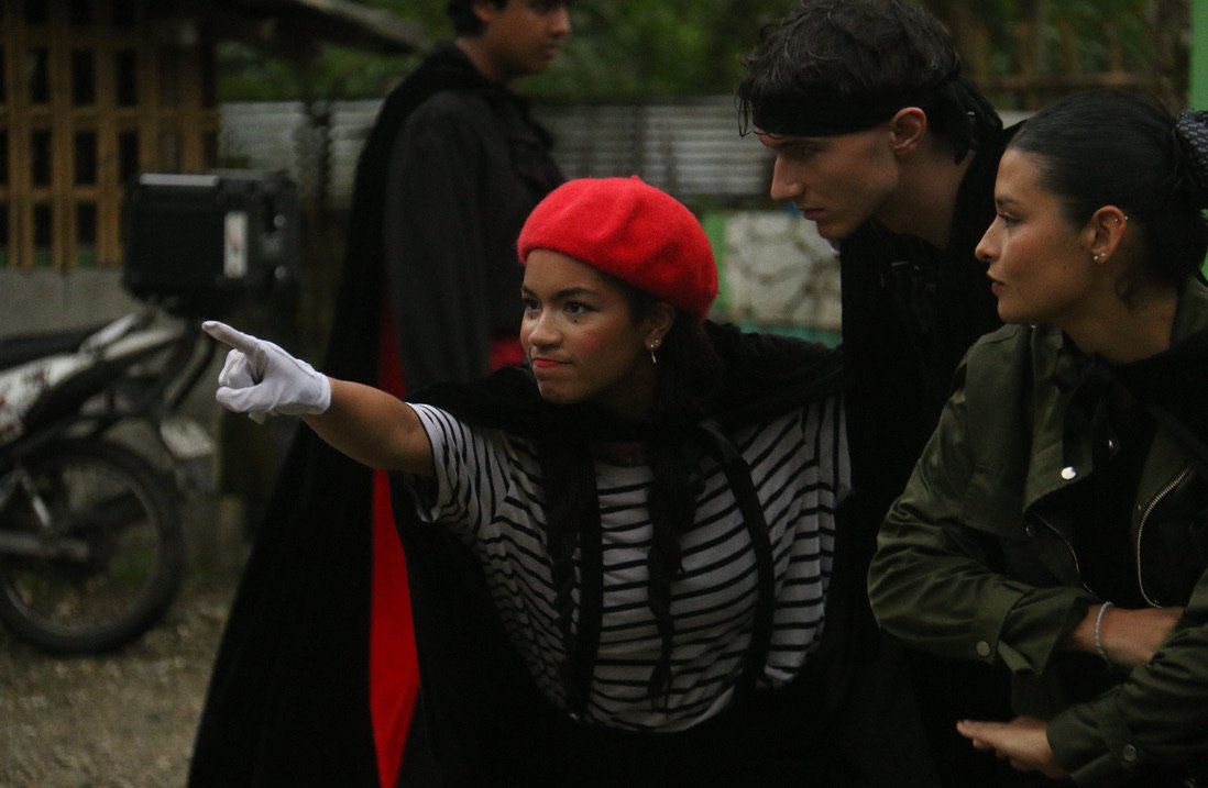 Three people outdoors, with one woman in a red beret and striped shirt pointing, two others looking at her.