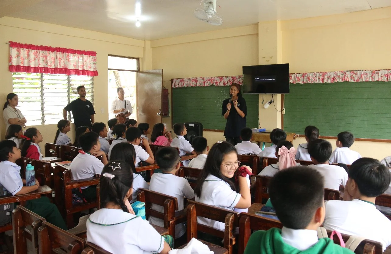 Students in a classroom listening to a woman speaking at the front, with some teachers standing near the windows and door, a television mounted on the wall, and a chalkboard behind the woman.