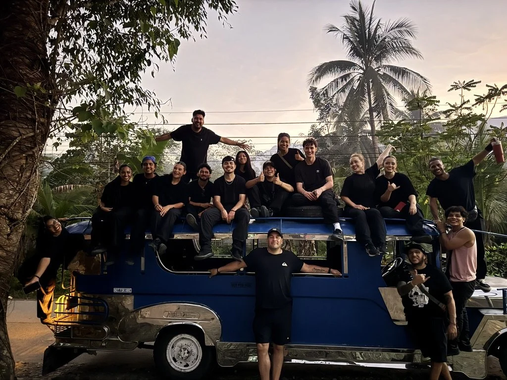 Group of people enjoying a ride on top of a blue and silver vehicle, with some sitting and others standing on the roof, surrounded by trees and palm trees with a sunset sky.