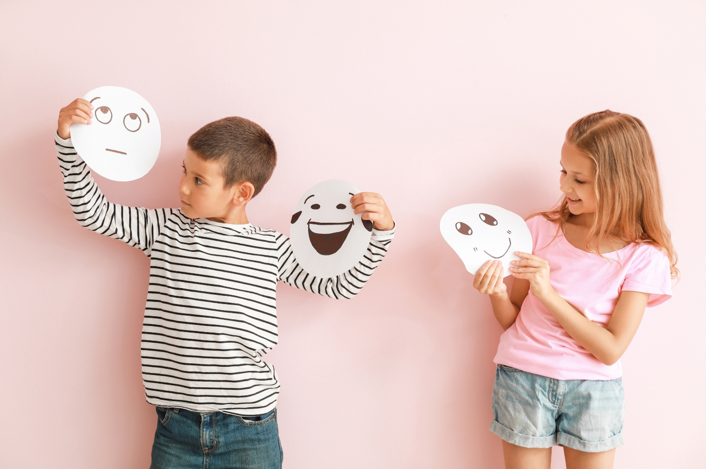 Zwei Kinder, ein Junge und ein Mädchen, halten Papiermasken mit lachenden und traurigen Gesichtsausdrücken vor einem pastelrosafarbenen Hintergrund.
