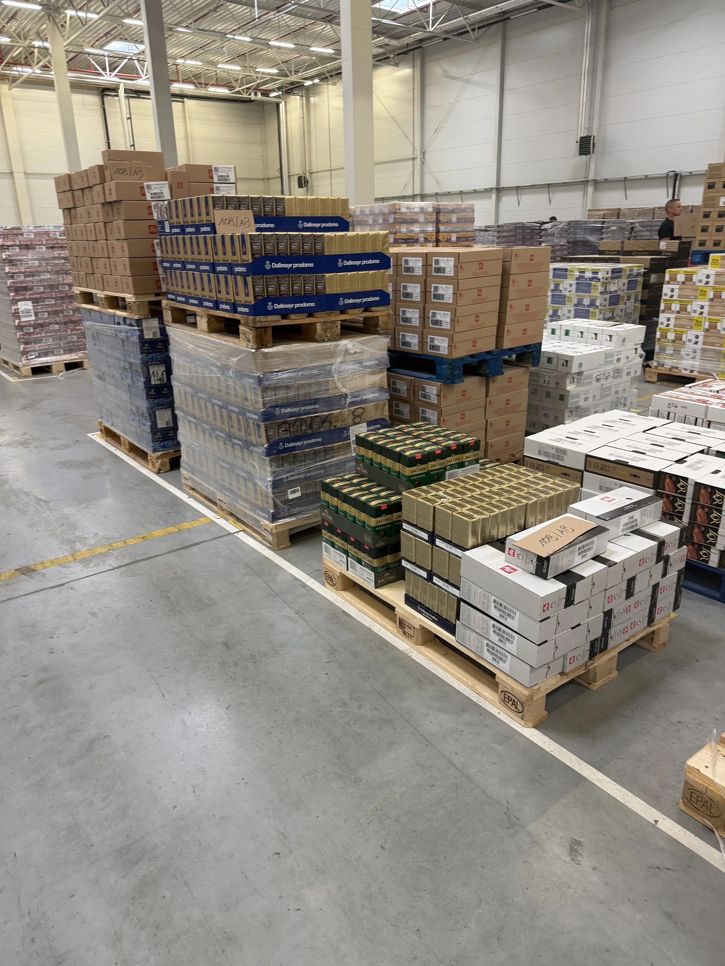 Warehouse with pallets of boxed goods stacked and organized, some wrapped in plastic, in a high-ceiling industrial space.