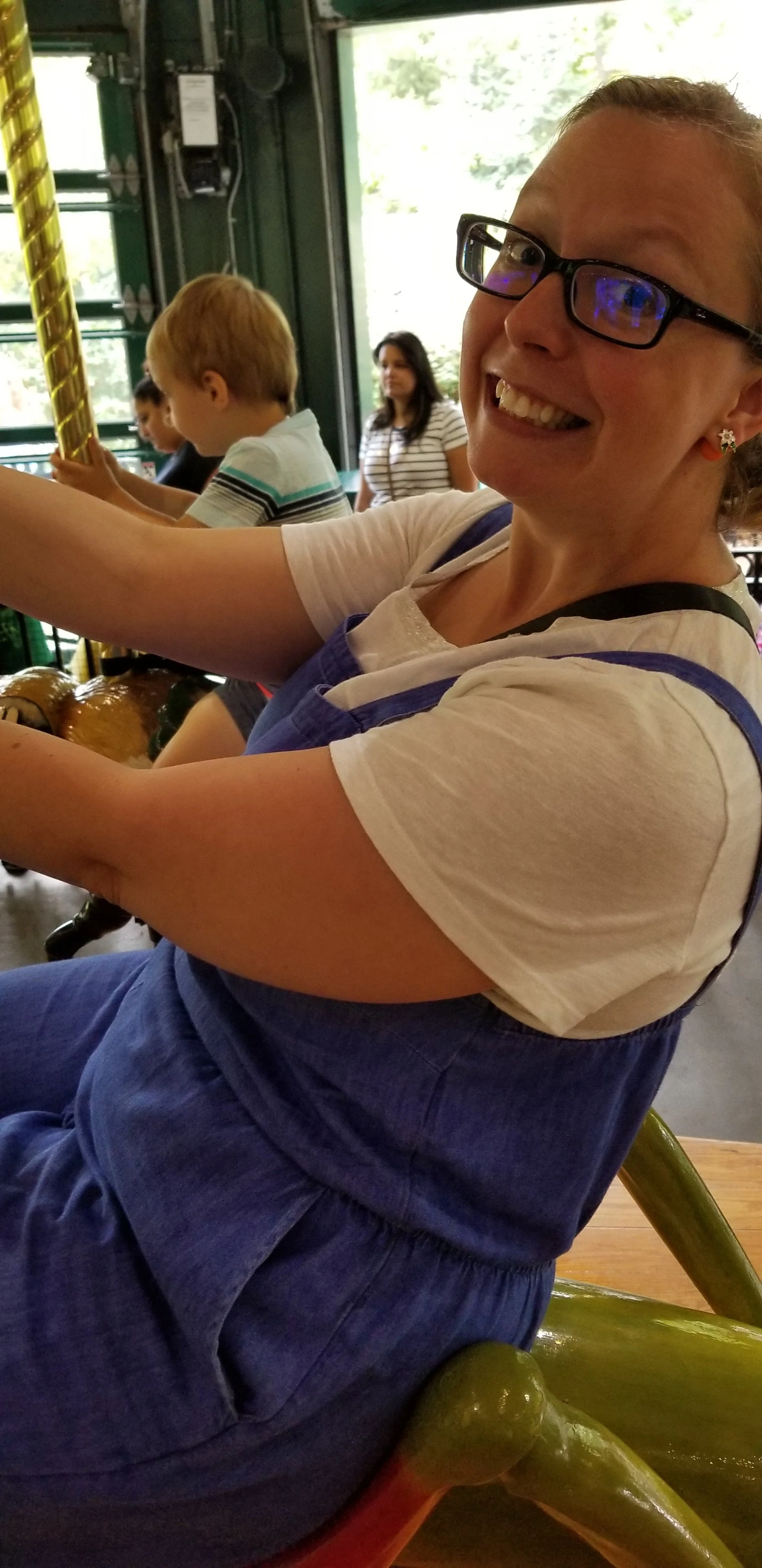 A woman smiling with glasses sitting on a green carousel horse at an amusement park, with children in the background.