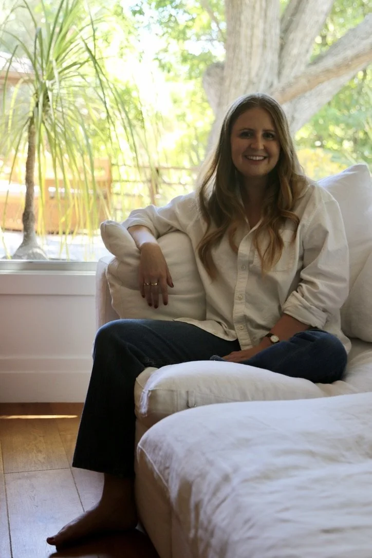 A woman with long, wavy hair sitting cross-legged on a white sofa, smiling, in a bright living room with large windows and outdoor greenery in the background.