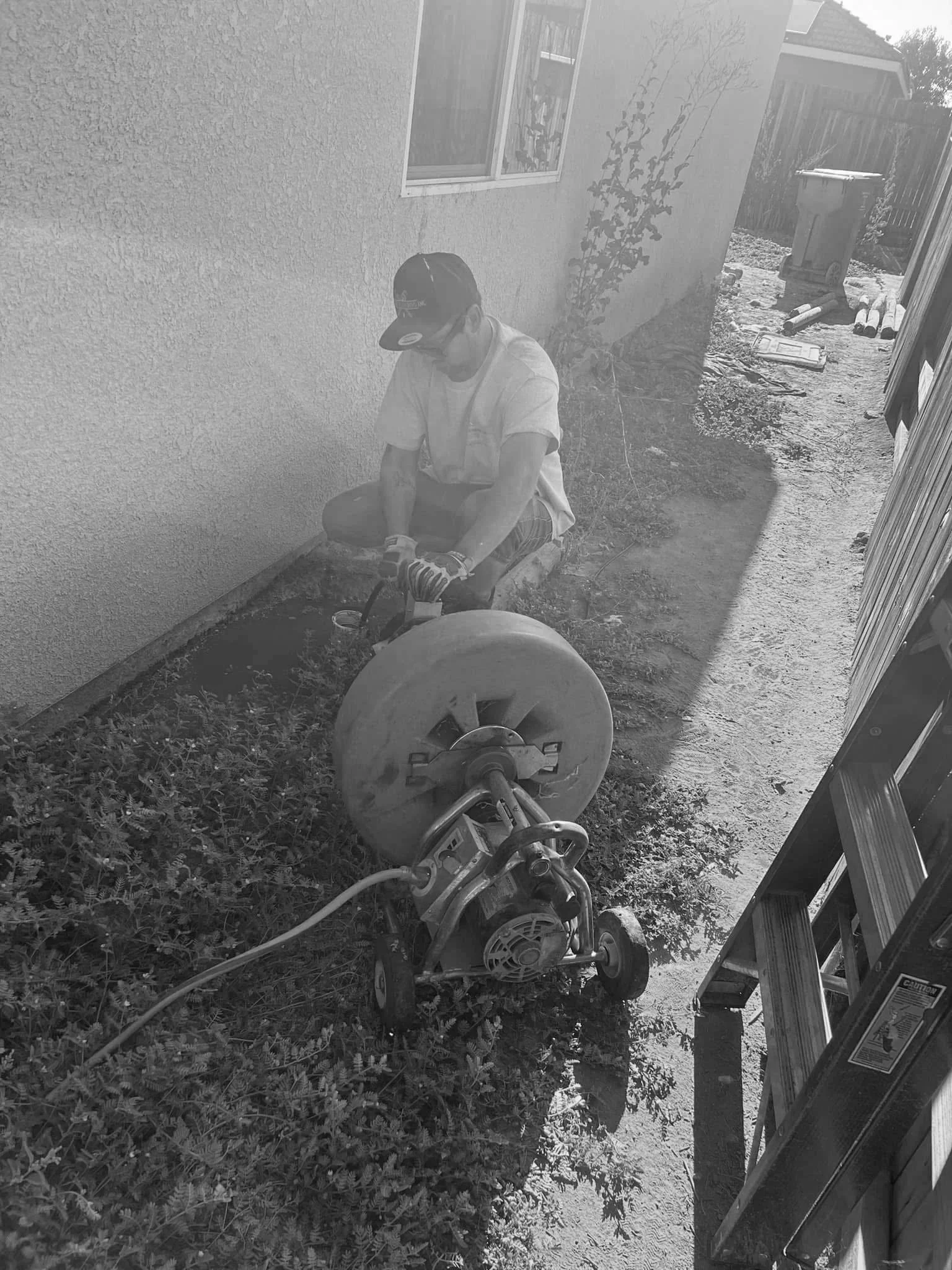 A person using a power saw outside a house, wearing safety gloves and a hat, with a trash bin and wooden fencing in the background.
