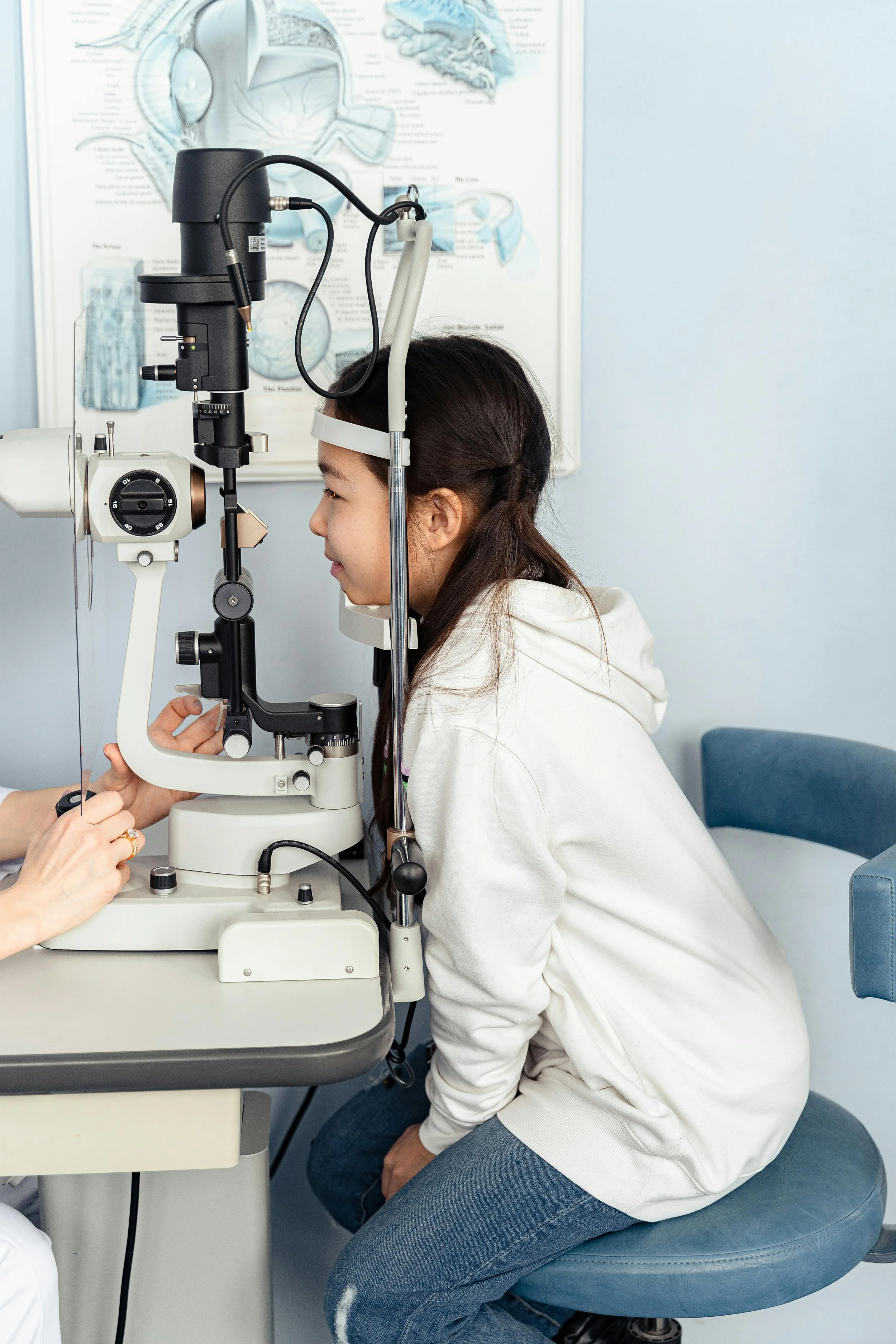 A woman undergoing an eye examination using a slit lamp microscope, in a front of an optometrist with anatomical charts in the background.